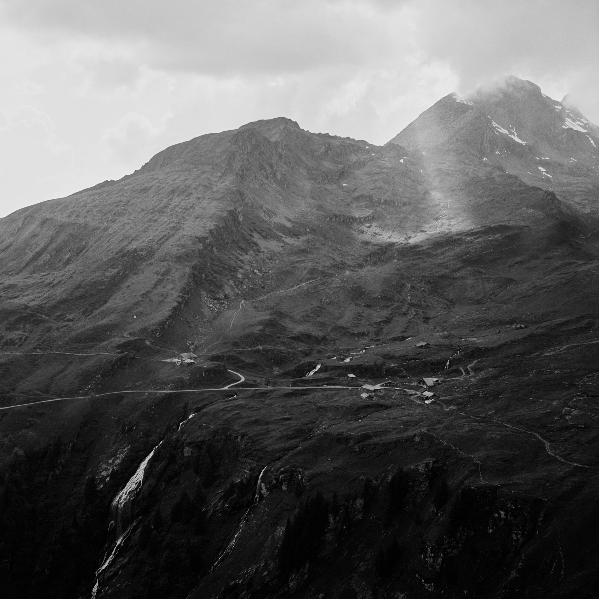 Black and white photo of a mountain landscape with steep slopes, small waterfalls, and scattered buildings at the base.
