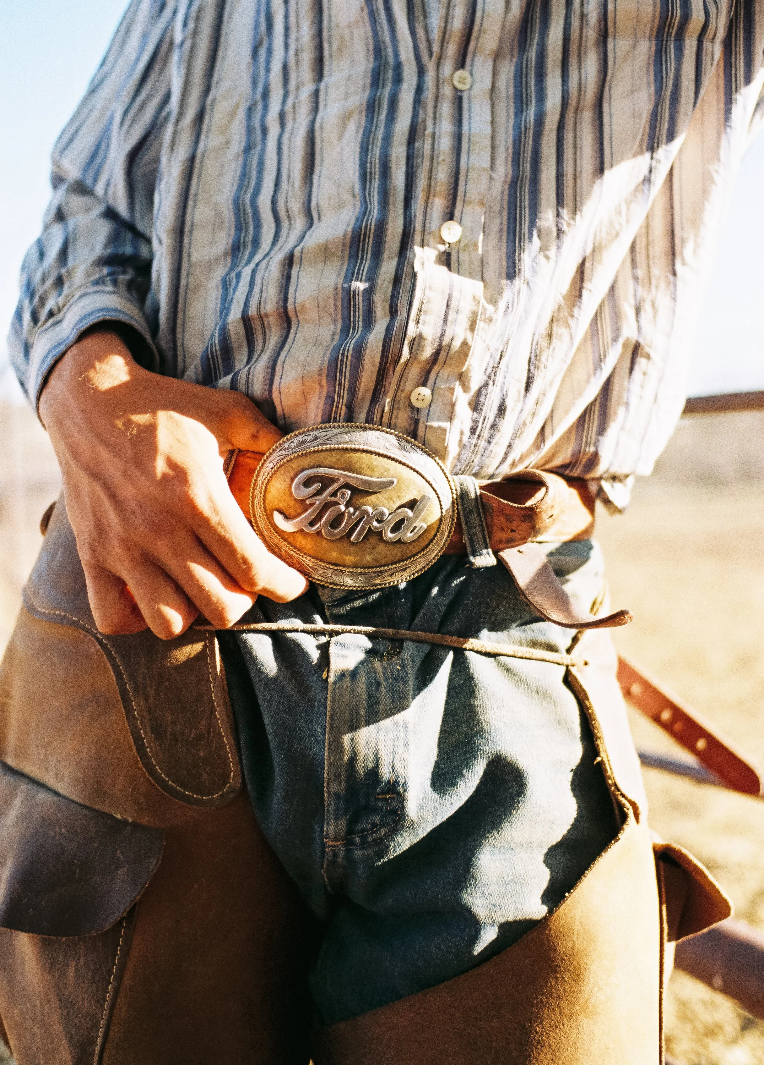 Close-up of a man wearing a striped shirt, jeans, and a leather belt with a large Ford logo buckle, standing outdoors.