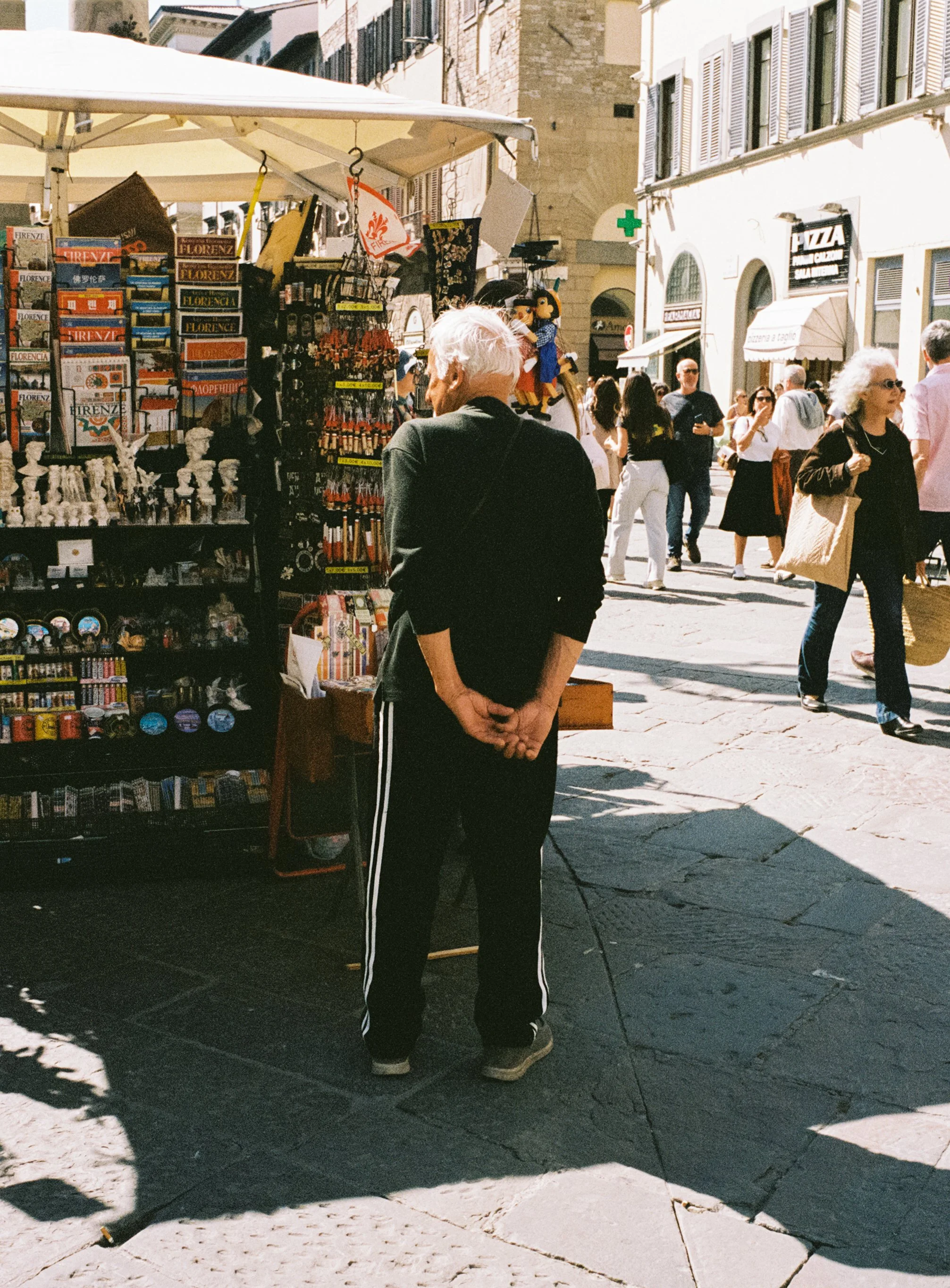 A street vendor stall in a busy outdoor market on a sunny day, with a man in black clothing looking at souvenirs like magnets and small figurines, and many people walking and shopping in the background.