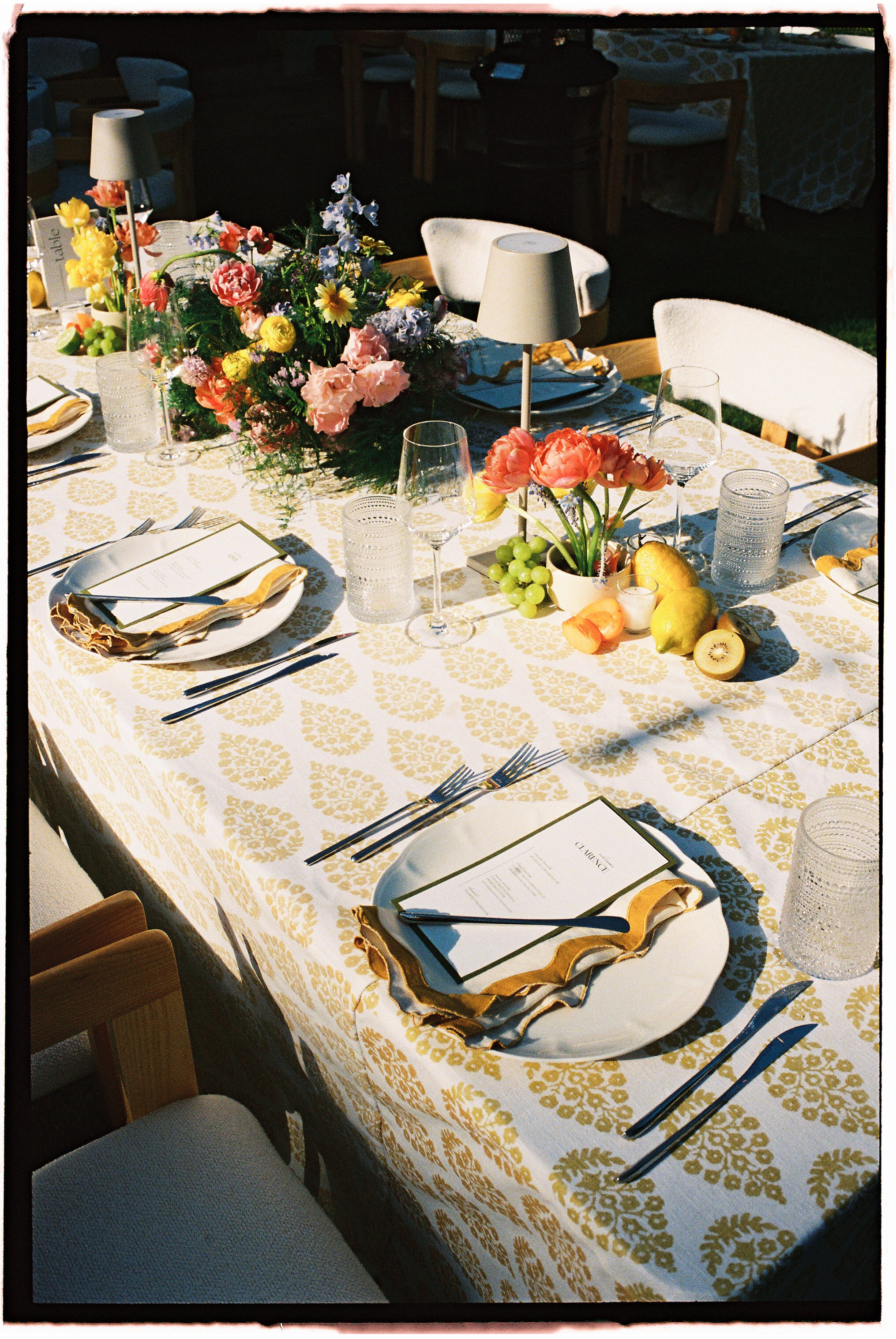 A decorated table set for a meal with a floral centerpiece, wine glasses, plates, utensils, candles, and fruit, in a well-lit outdoor setting.