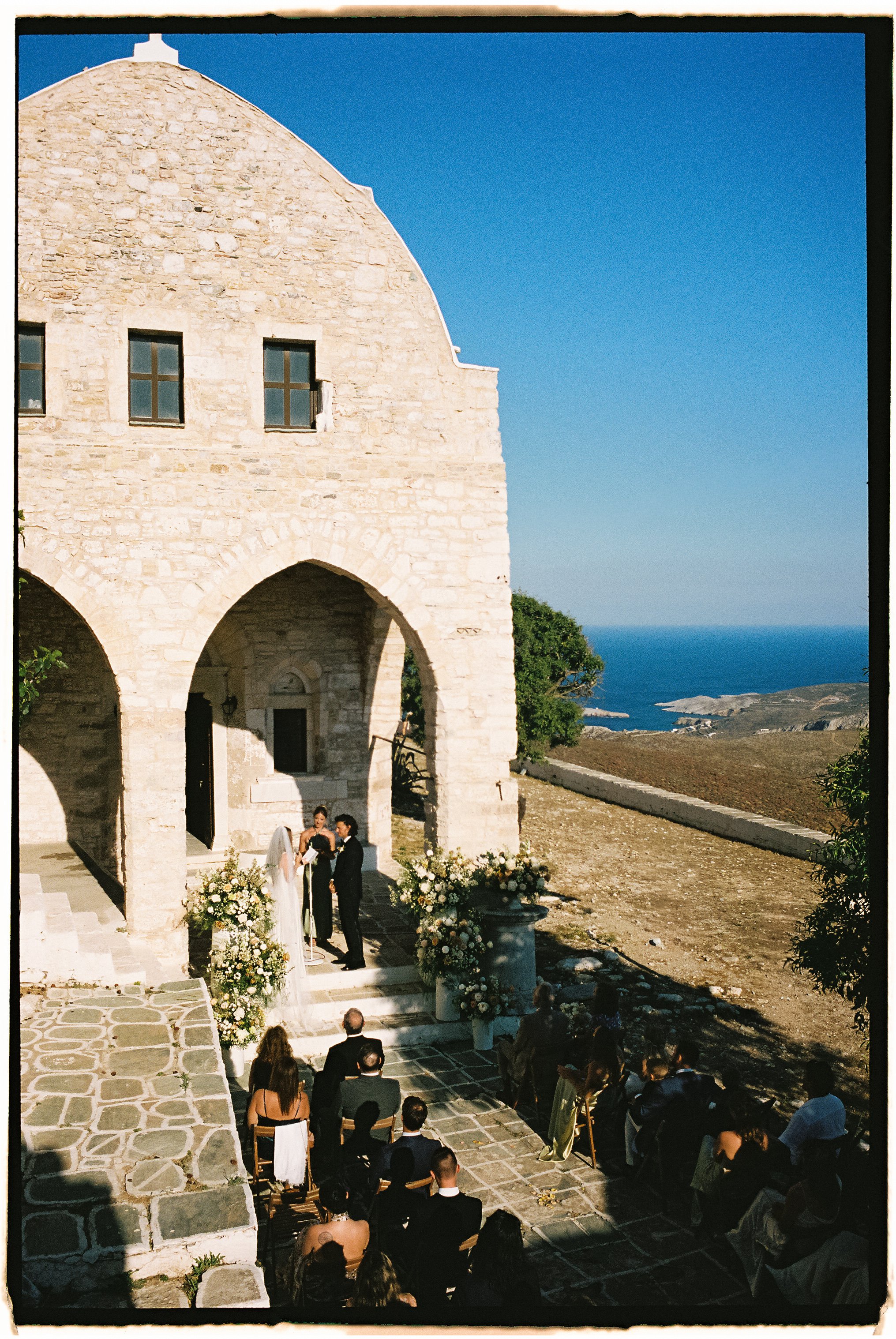 A wedding ceremony taking place outdoors in front of a stone building with arches. The couple is standing under an arch decorated with flowers, with a minister officiating. Guests are seated on chairs in front of the couple, and the backdrop features