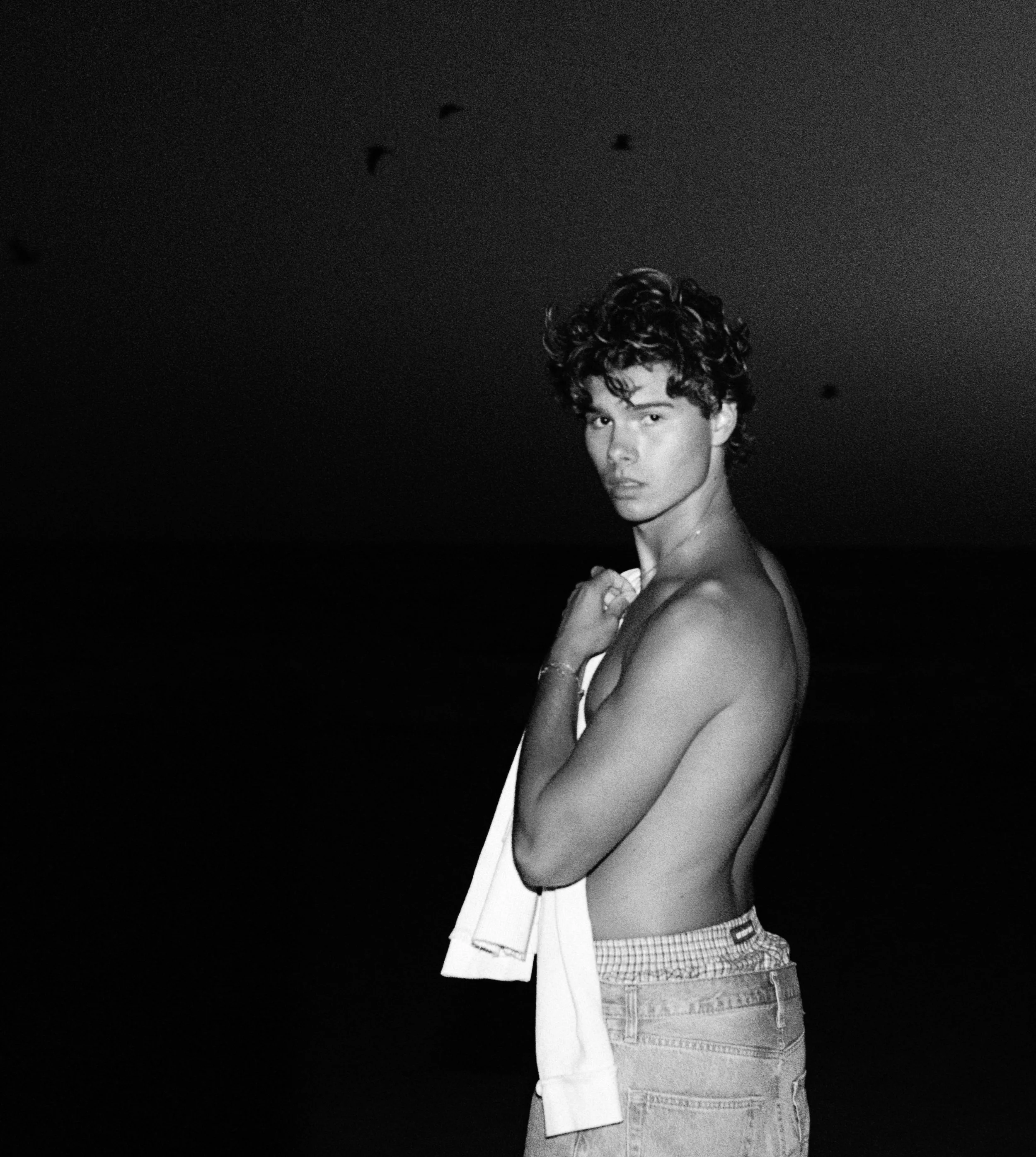 A young man with curly hair standing shirtless at the beach at night, holding a towel over his shoulder, facing the camera.
