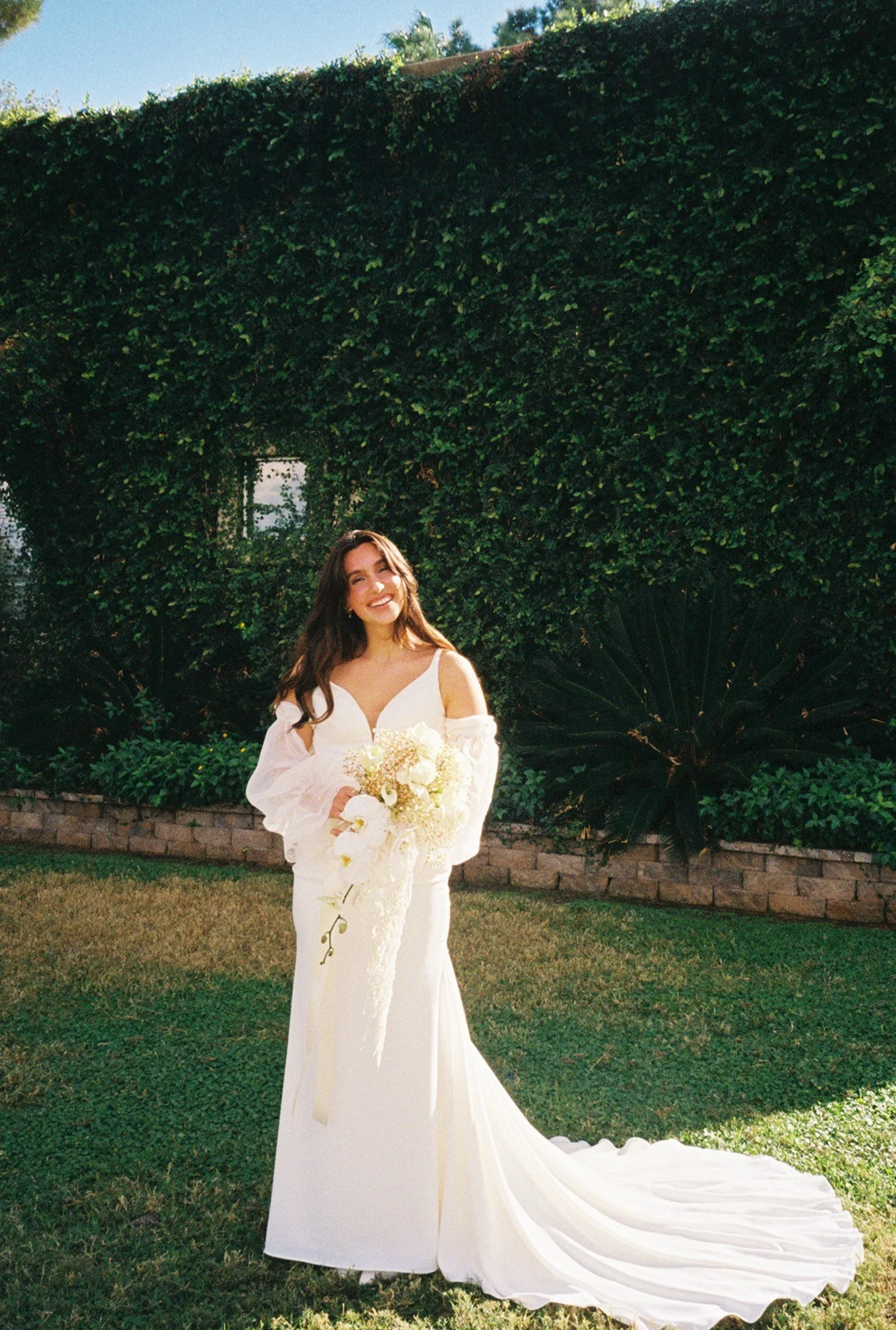 A woman in a white wedding dress holding a bouquet of flowers, standing on grass in front of green foliage and a brick border.