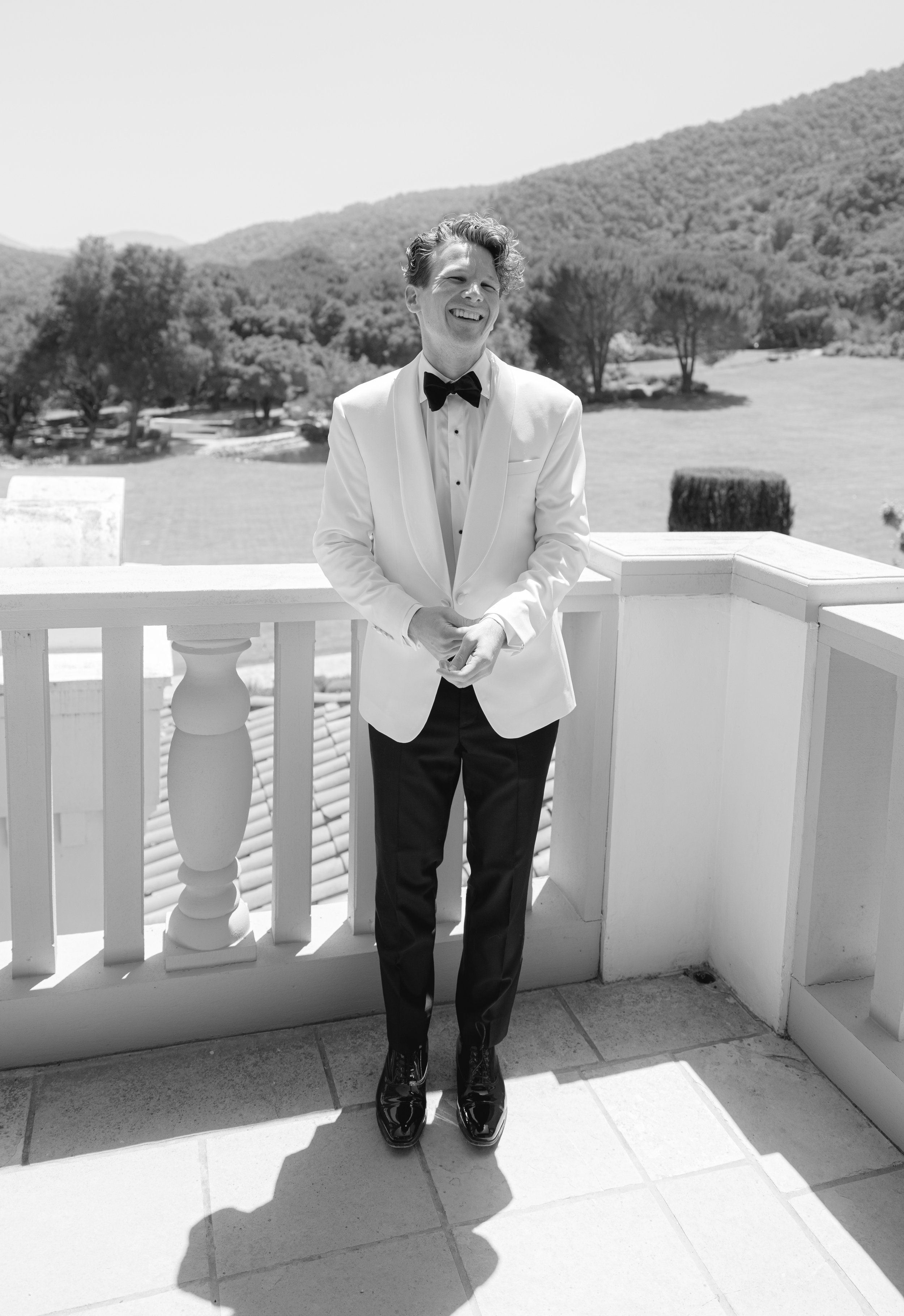 A young man in a tuxedo with a bow tie, smiling and standing by a white railing on a terrace with a scenic background of trees and mountains.