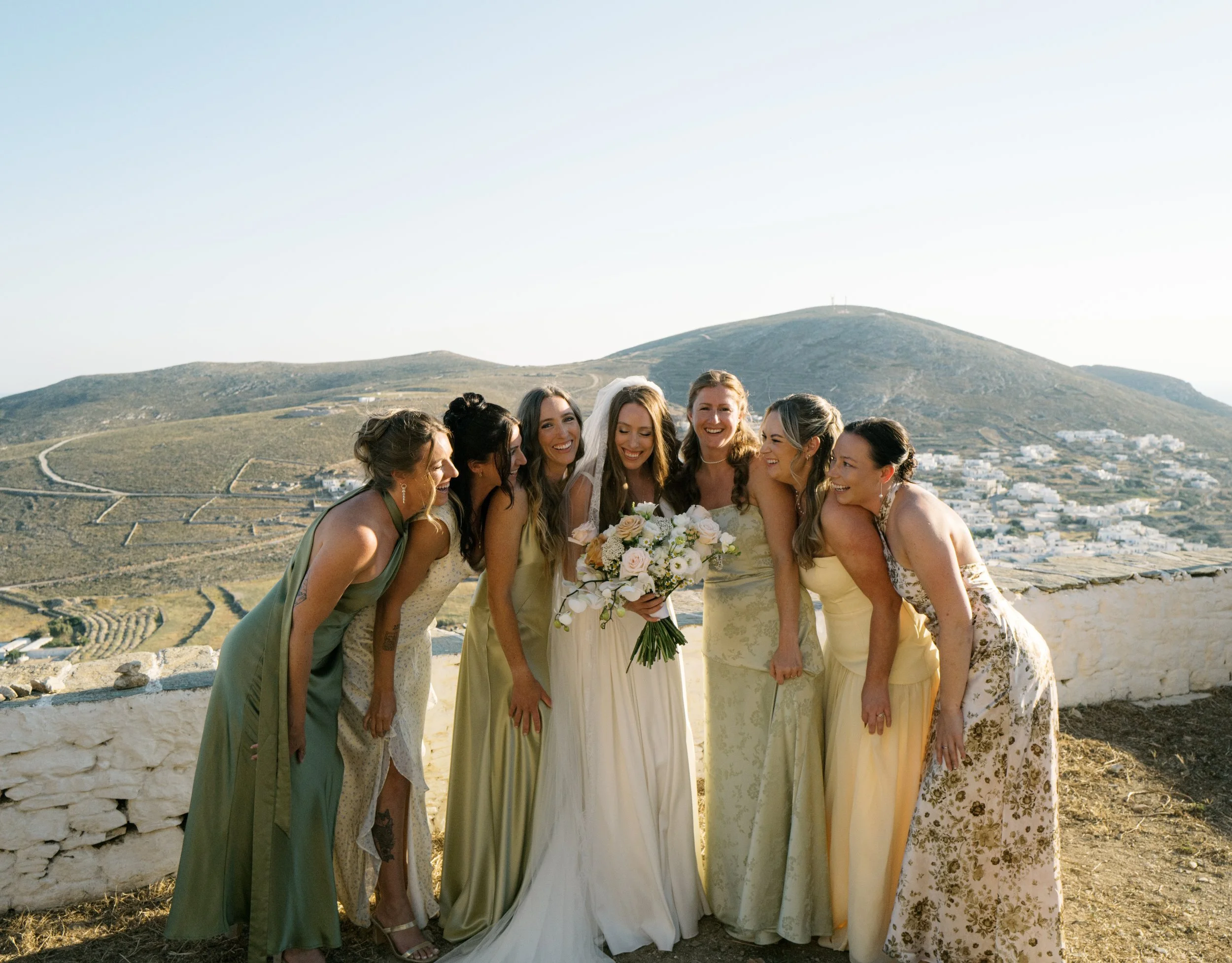Group of seven women in dresses, smiling and laughing with a scenic hillside and white buildings in the background, during a daytime outdoor event.