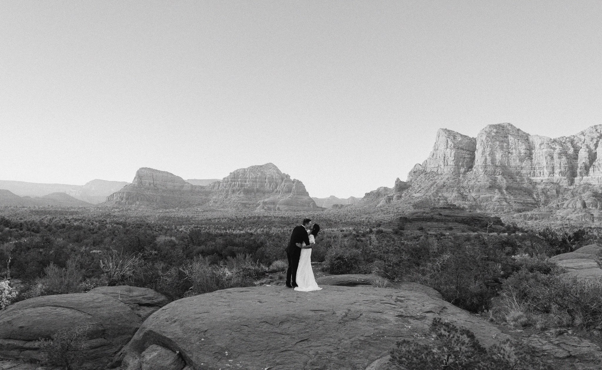 A couple in wedding attire standing in a scenic desert landscape with large rock formations in the background, embracing and gazing into each other's eyes.