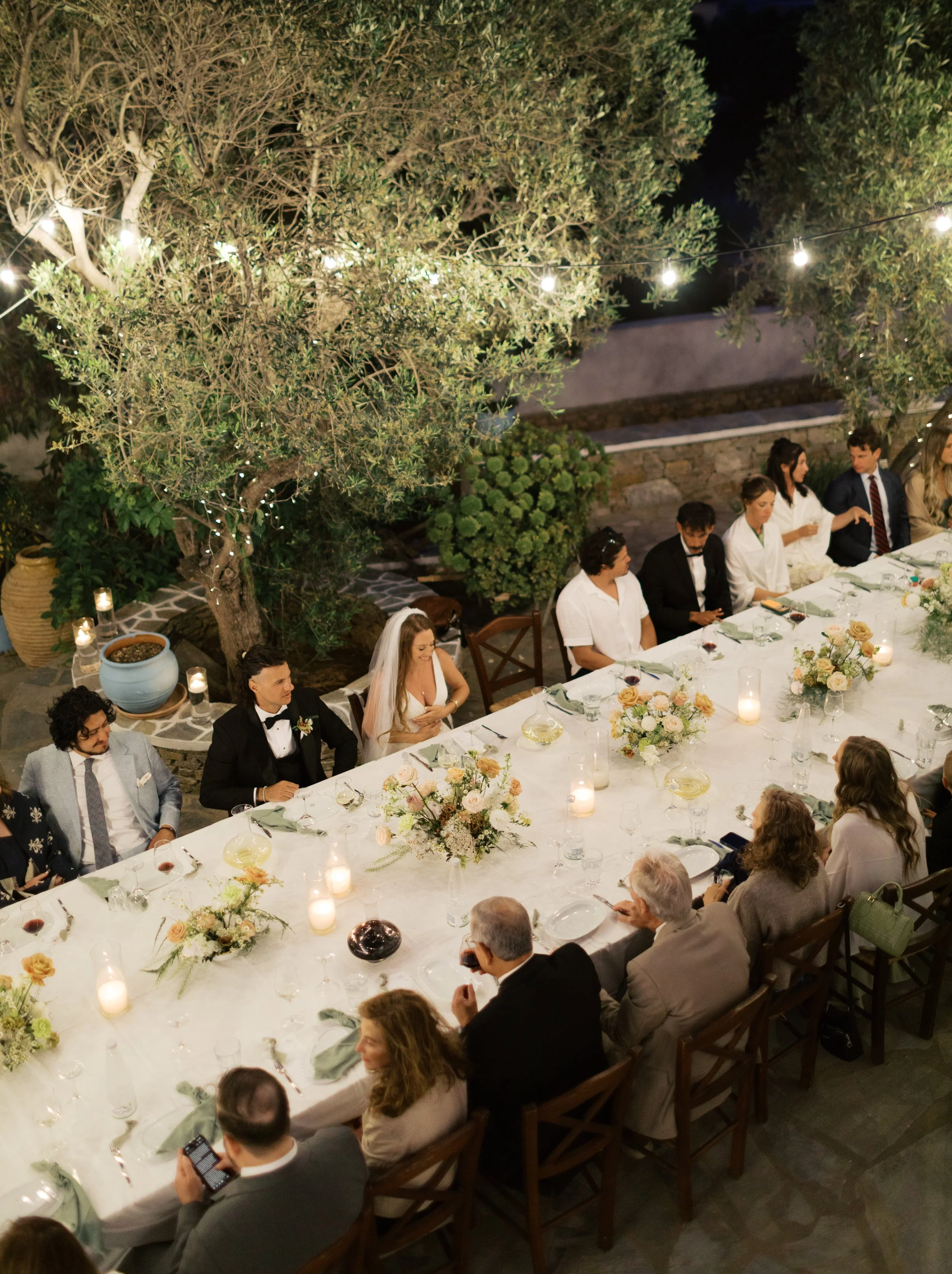 A nighttime outdoor dinner party with guests sitting around a long table decorated with floral centerpieces and candles, under string lights and trees.