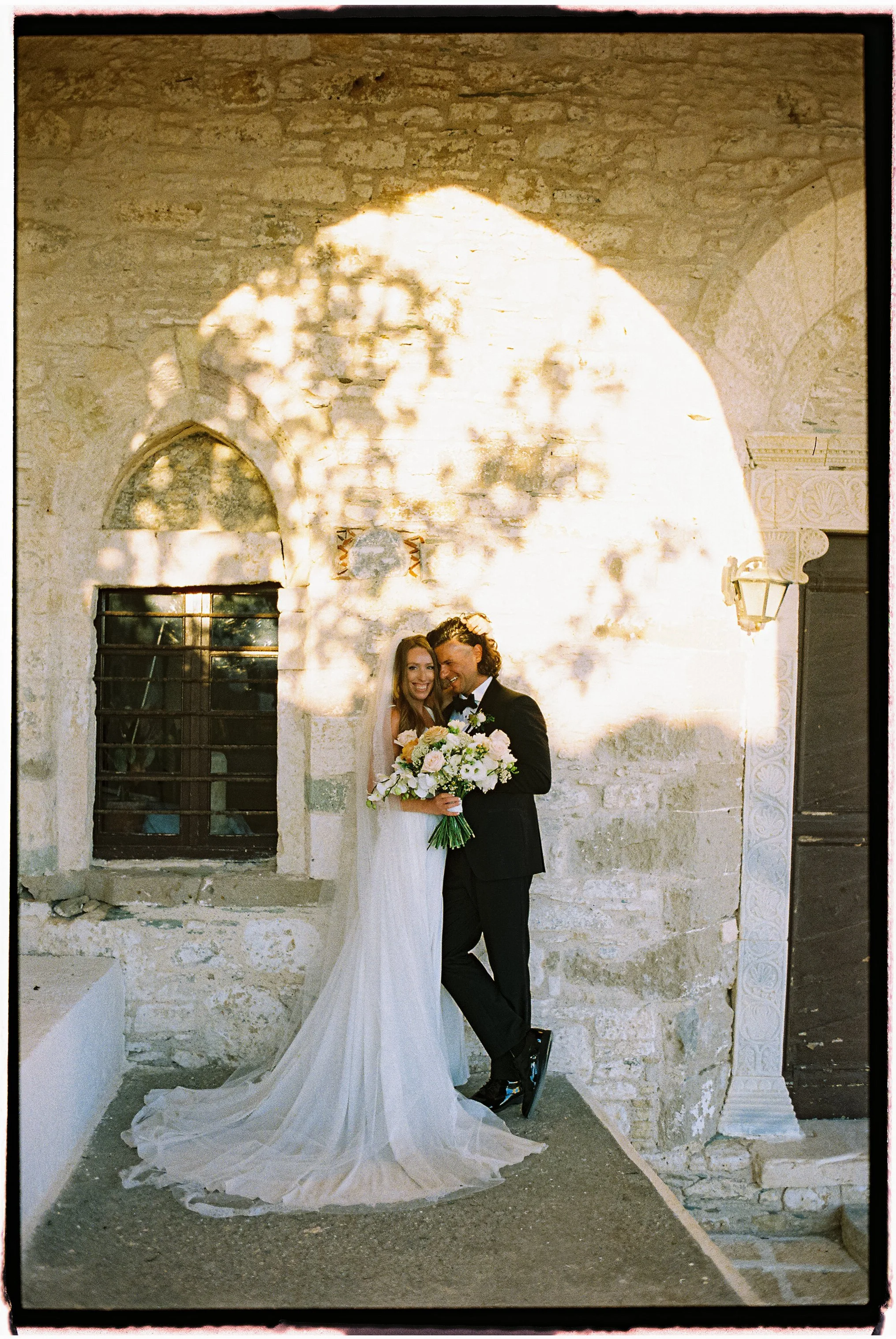 A bride and groom pose in front of a rustic stone wall at their wedding. The bride wears a white wedding gown with a long train, and the groom is dressed in a black tuxedo. They are smiling and holding a bouquet of white and pink flowers.