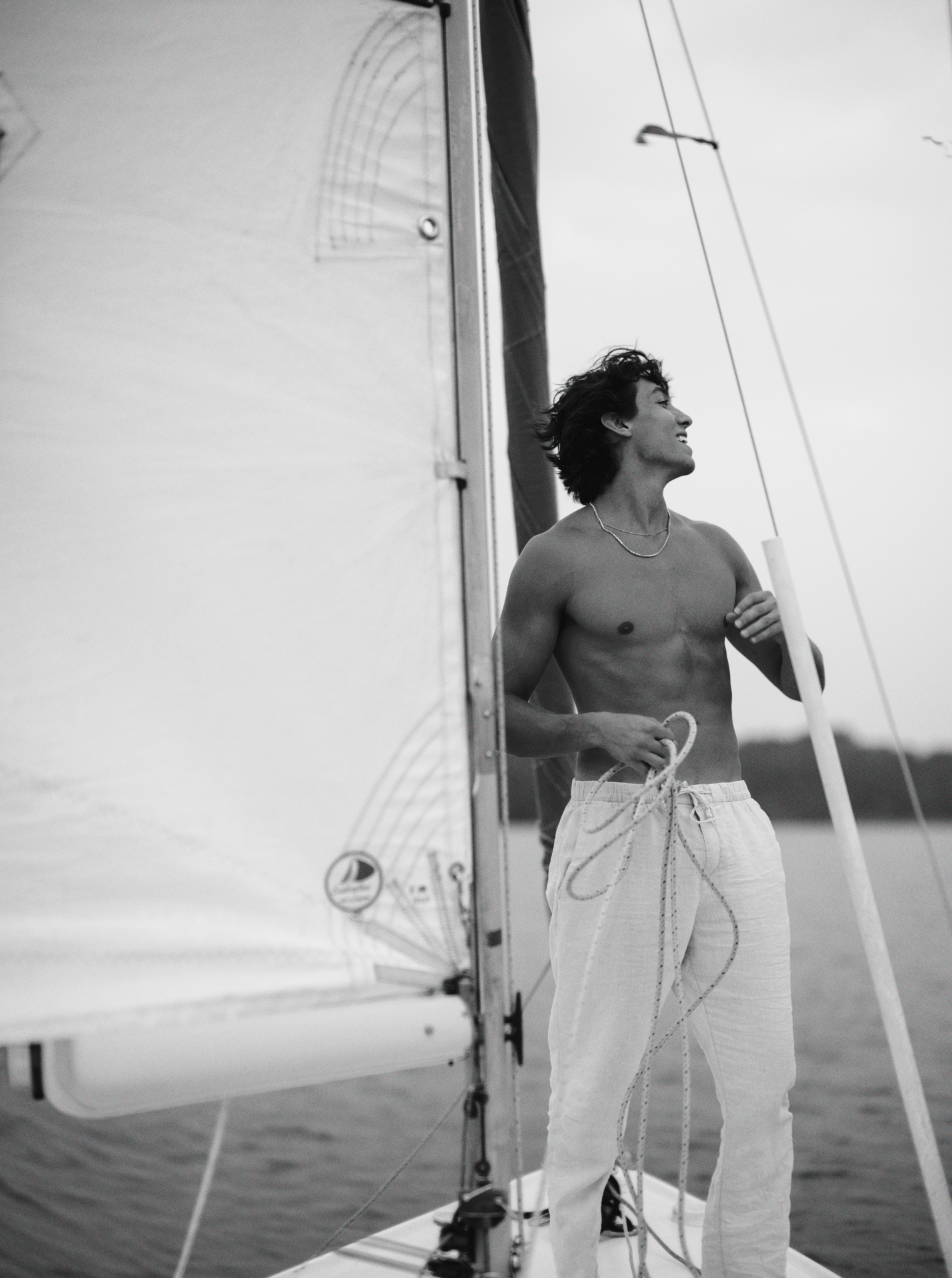 A shirtless young man with dark, wavy hair standing on a sailboat, holding a rope in one hand and smiling while looking to the right. He is wearing light-colored, loose-fitting pants and a necklace, with water and a cloudy sky in the background.