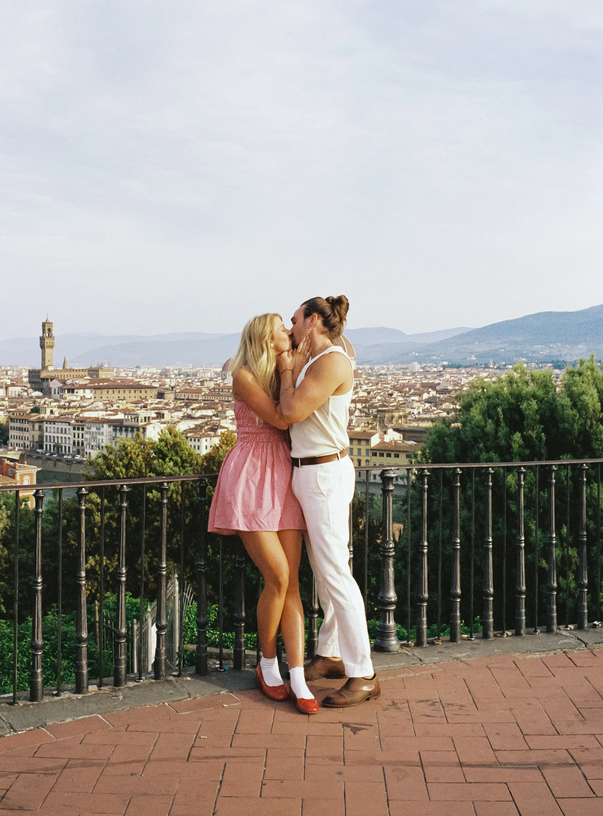 A couple sharing a kiss on a balcony overlooking a cityscape with mountains in the background.