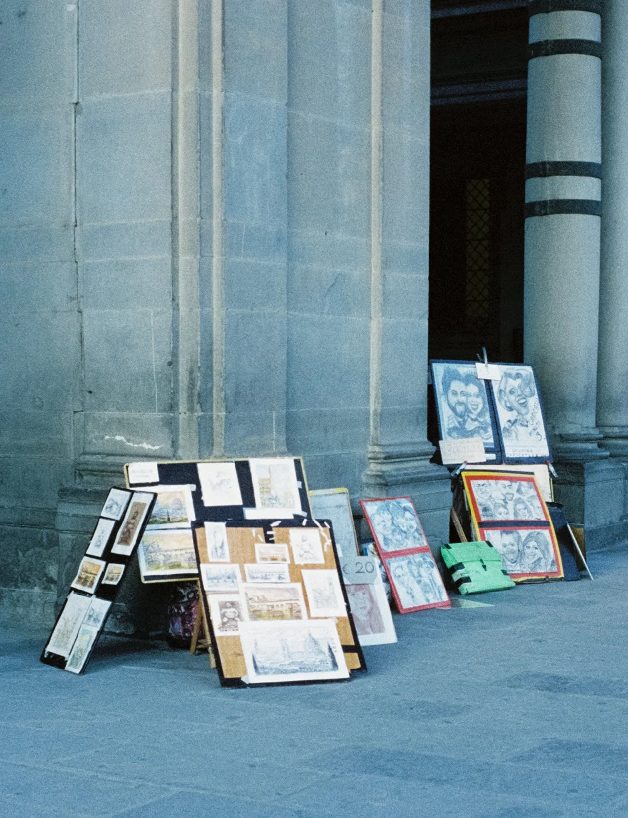 Street art sale display featuring caricature portraits on framed boards against a stone building wall.