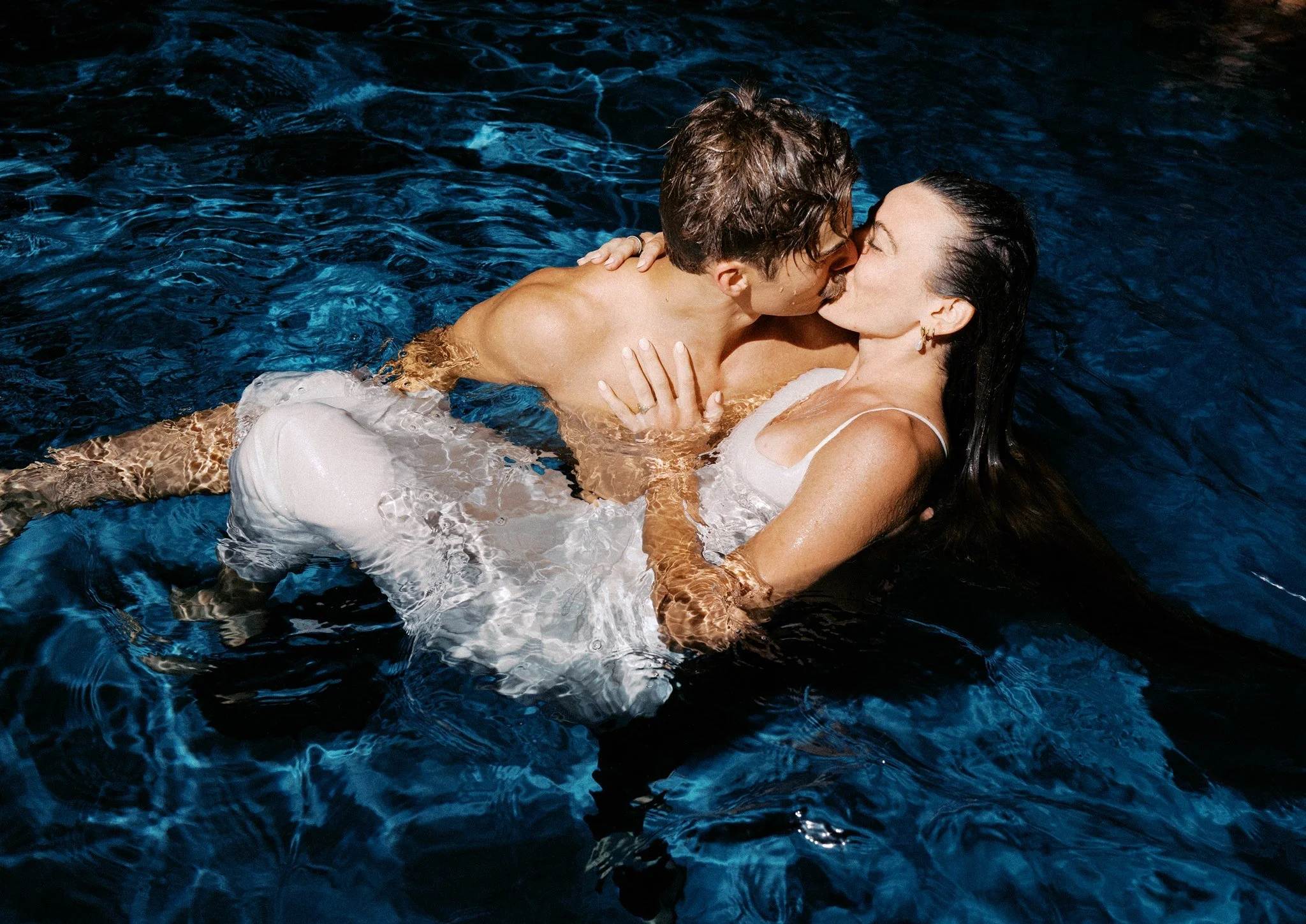 A man and woman are cuddling and kissing in a pool, surrounded by water with ripples.