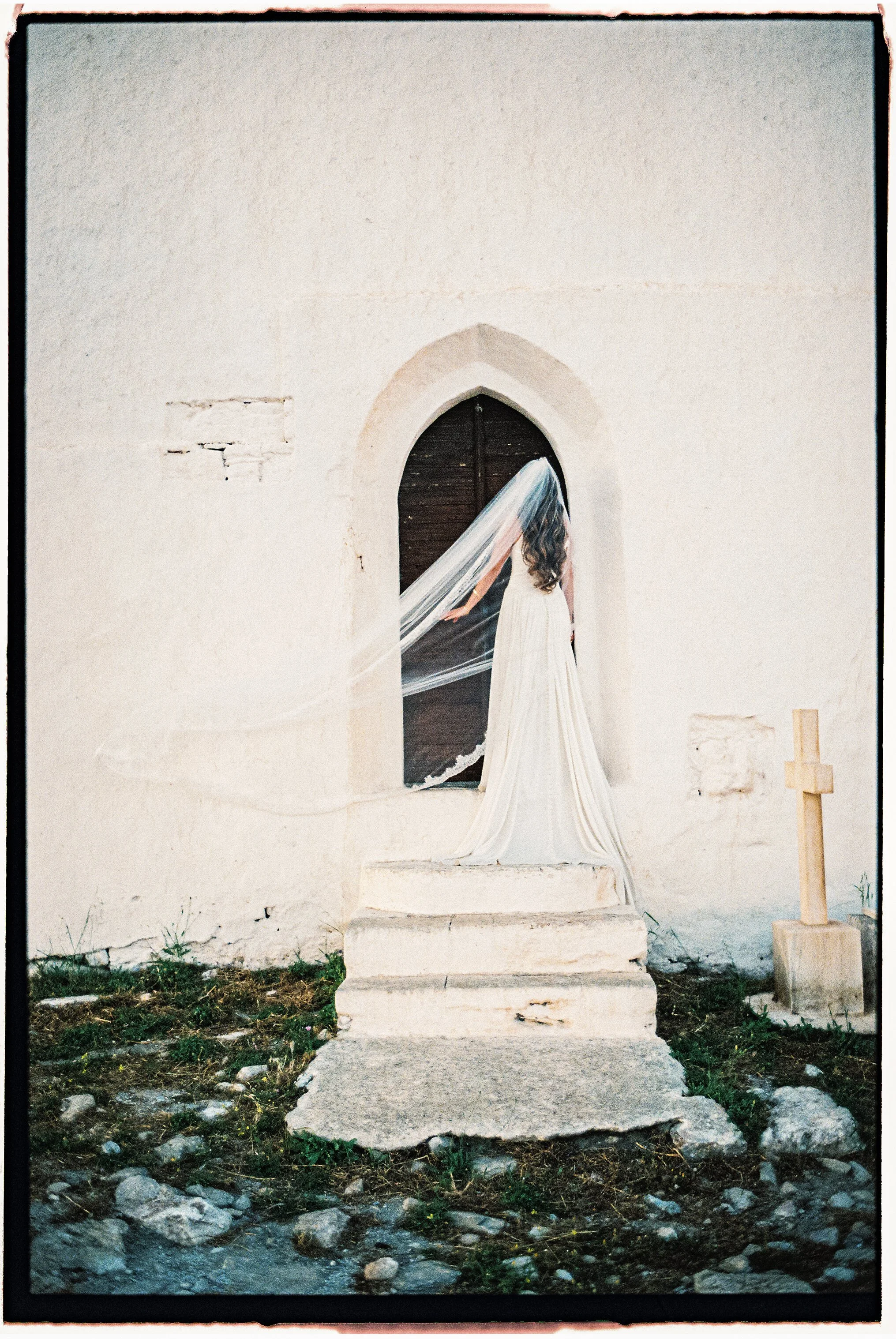 A woman in a white wedding dress with a veil standing at an open wooden door in a white stone building with steps leading up to it, a cross on the ground to the right of the steps, and a gravel pathway in the foreground.