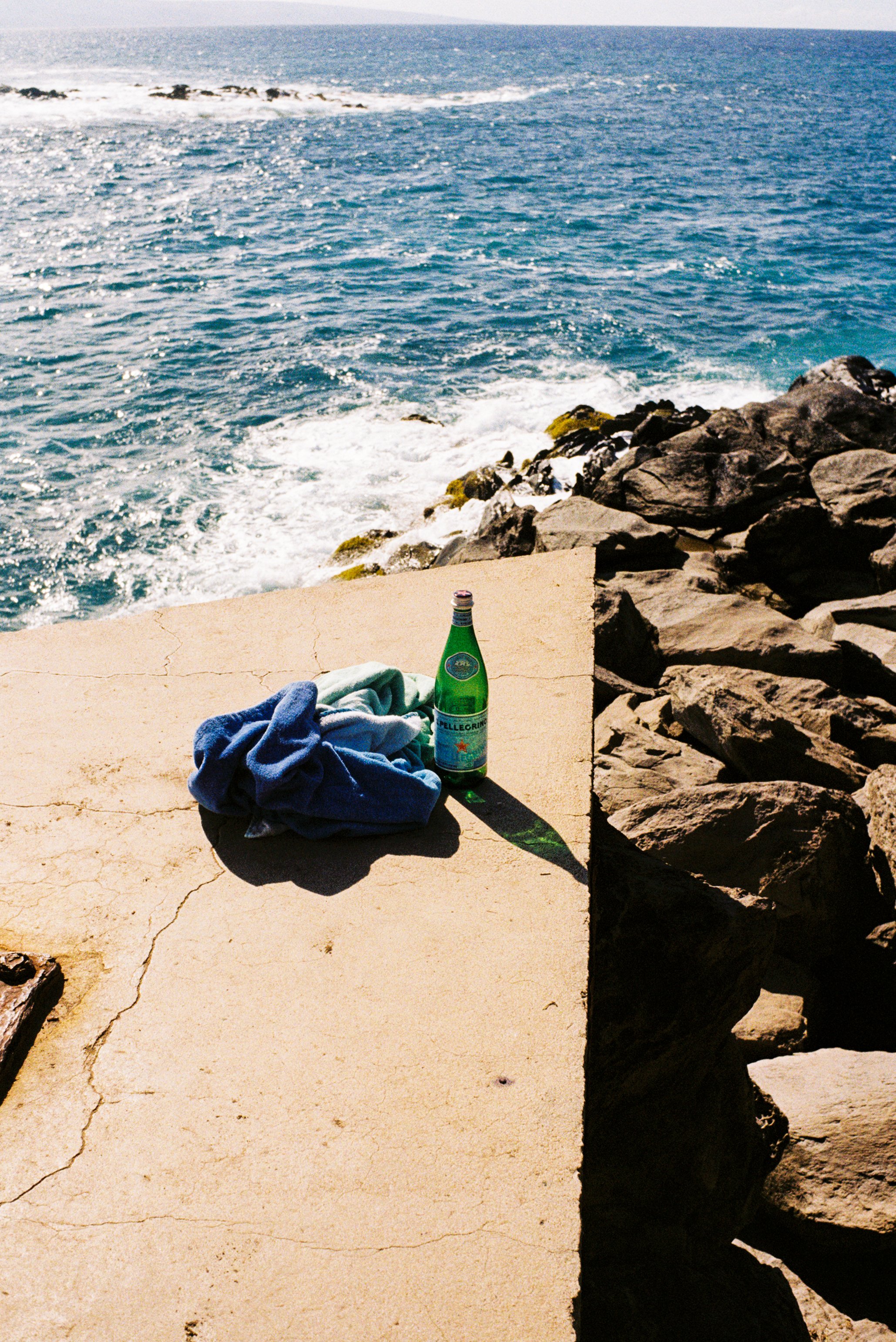 A green glass bottle of Pellegrino sparkling water, a blue towel, and a green towel on a cracked concrete surface near the water, with rocks and ocean waves in the background.