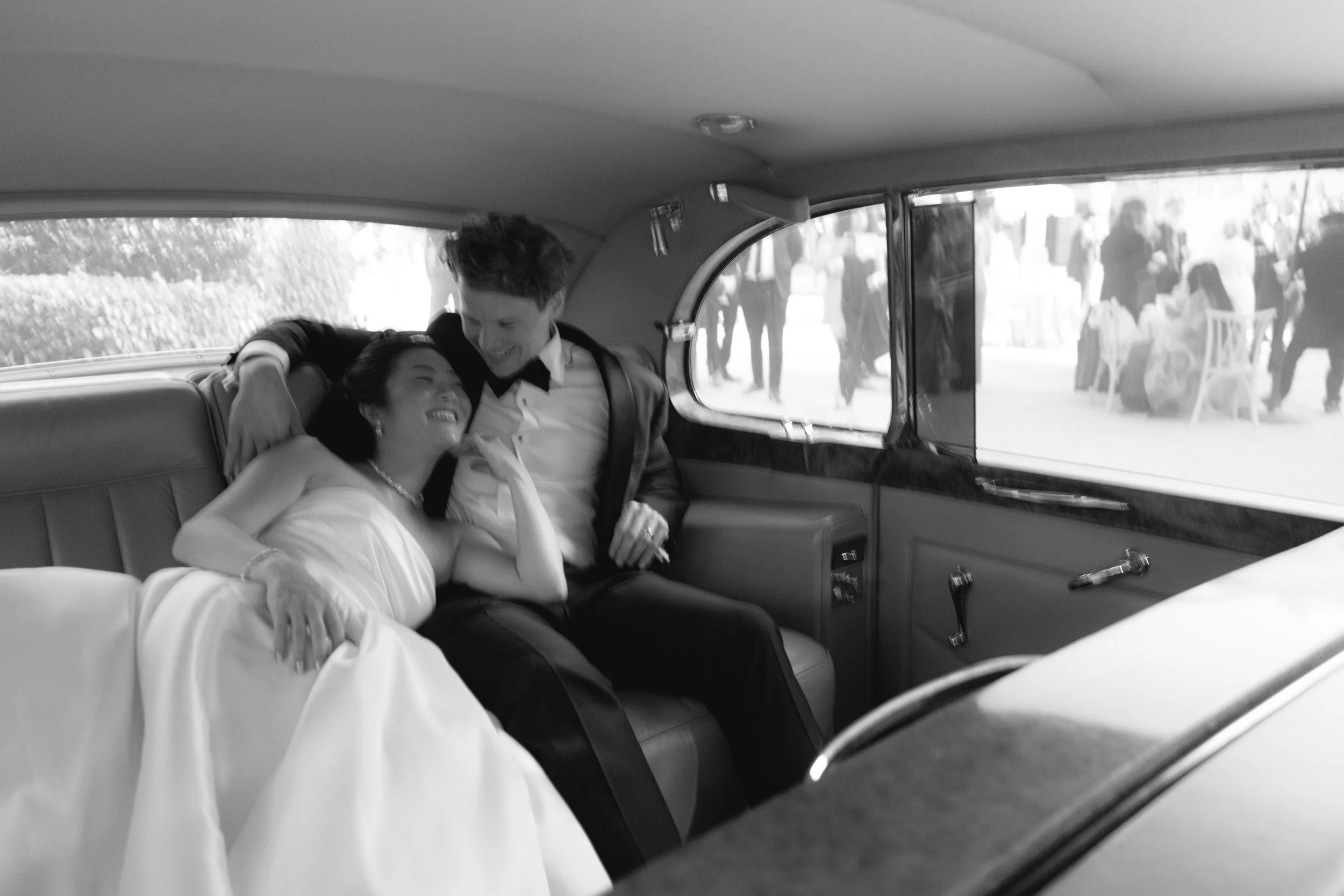 A bride and groom sitting inside a vintage car, embracing and smiling at each other, with people gathered outside.