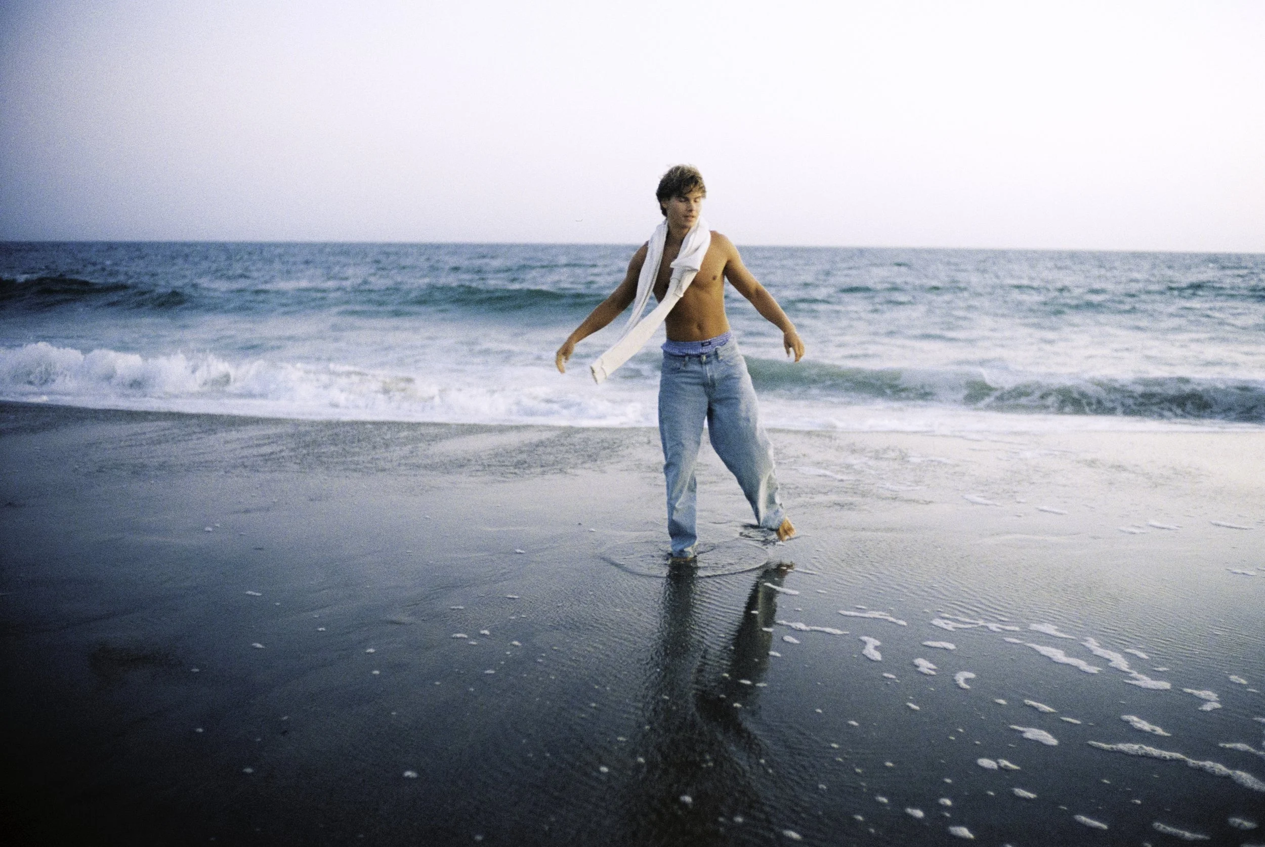 A shirtless young man with a white scarf around his neck, wearing jeans, walking barefoot on the wet sand at the beach near the ocean with gentle waves.