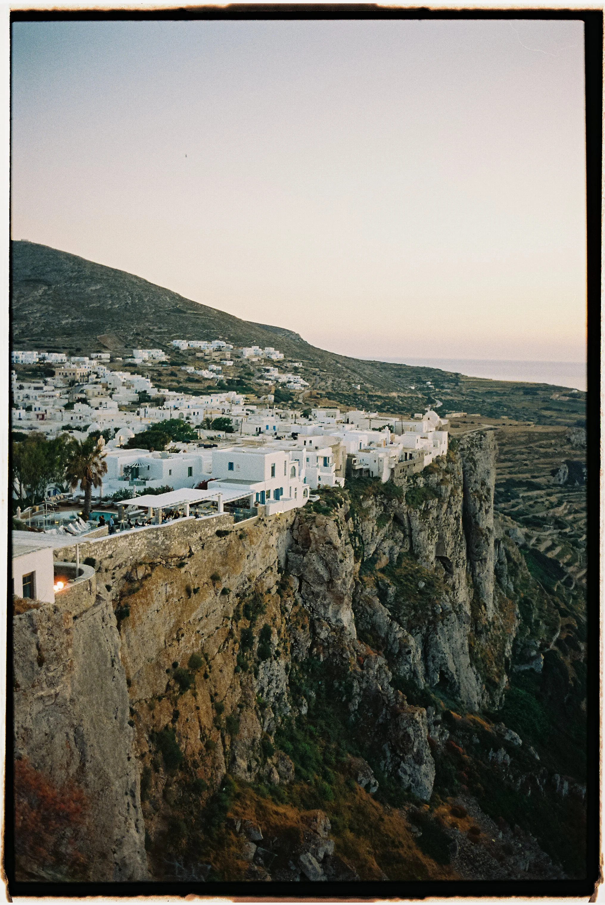 Scenic view of a cliffside village with white buildings, blue accents, and outdoor seating areas overlooking the sea at sunset.