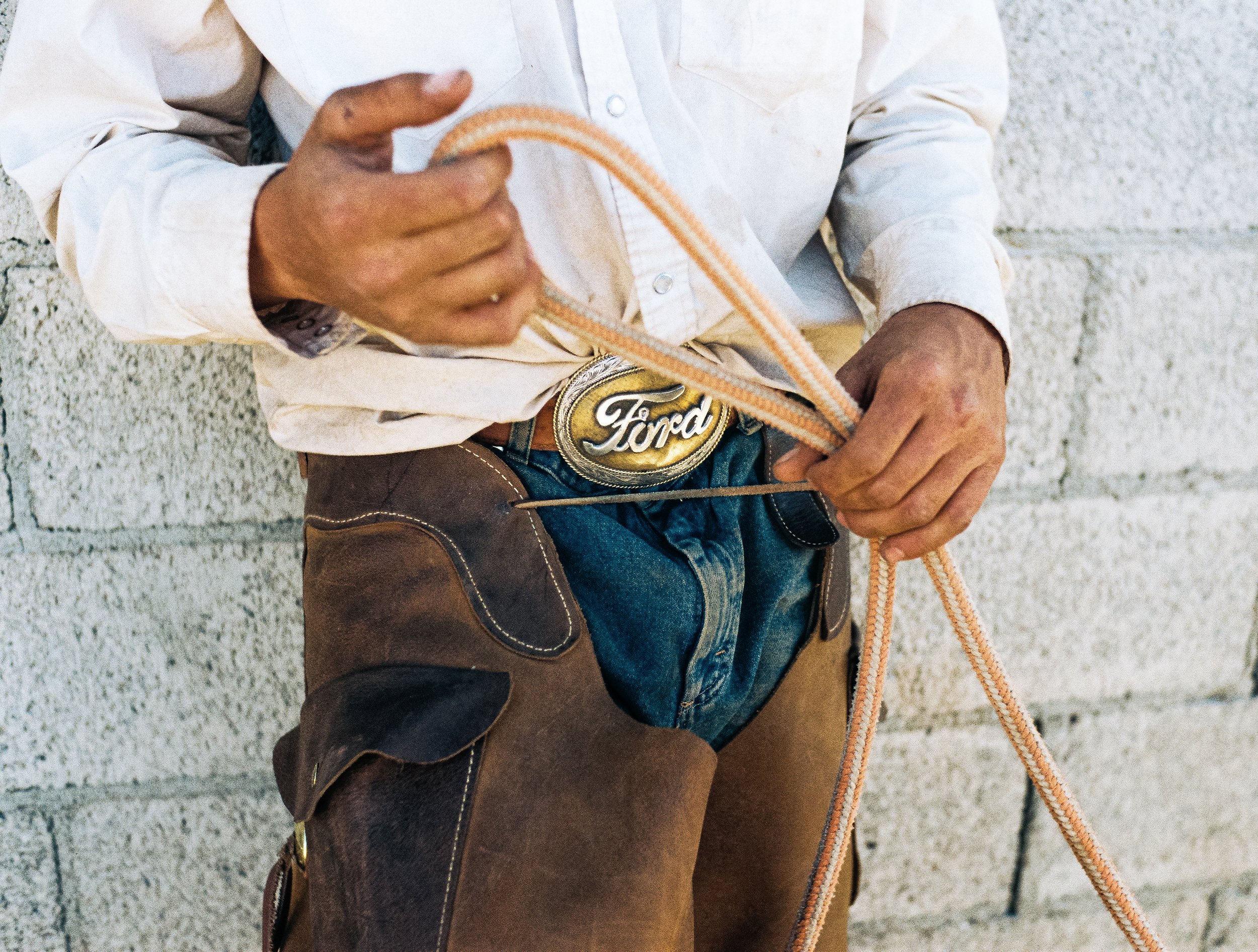 A man dressed as a cowboy, wearing a white shirt, leather chaps, and a large belt buckle with the Ford logo, holding a lasso in front of a brick wall.