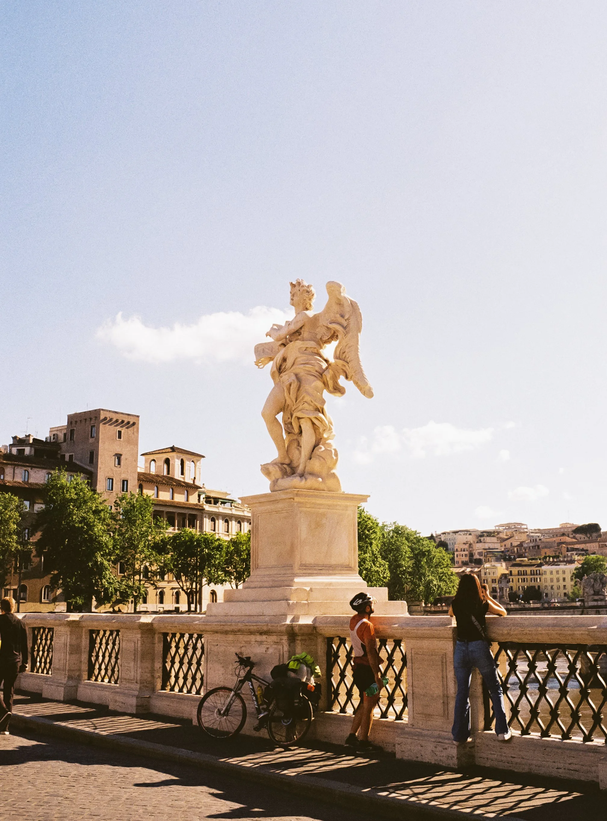 Statue of a figure holding a fish, located on a bridge with people and a bicycle nearby, overlooking a cityscape with trees and buildings.