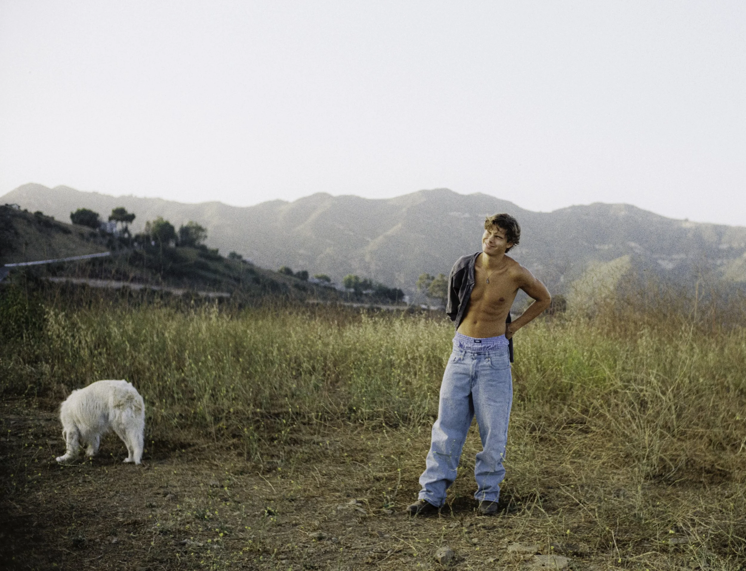 A shirtless young man with light brown hair stands in a field, holding a gray jacket over his shoulder, with a small white dog nearby grazing, and hills in the background under a clear sky.