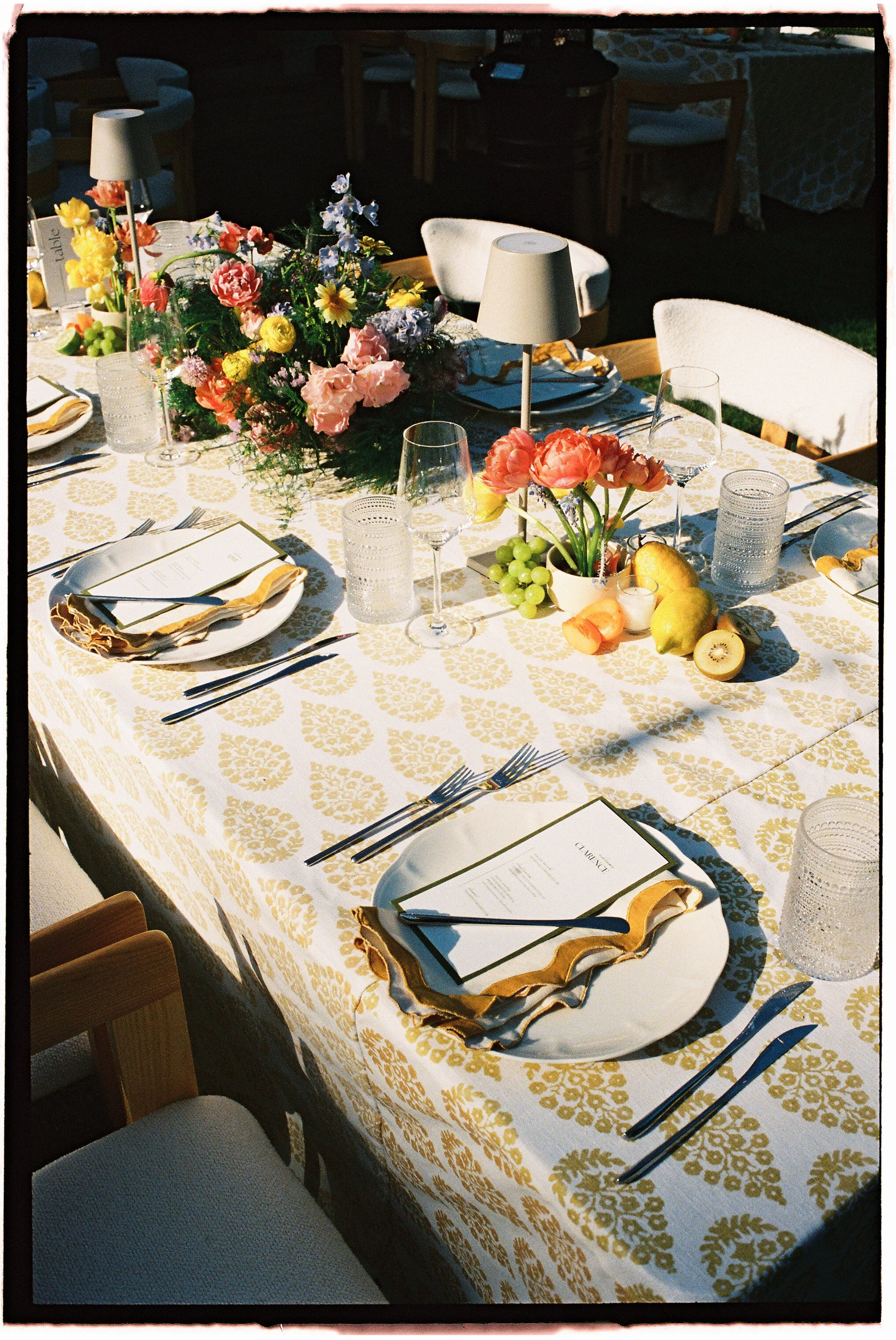 A decorated dining table set for a meal with plates, utensils, glasses, and gold and white napkins. The centerpiece consists of colorful flowers in a vase, and a smaller arrangement of pink flowers with green grapes and fruit around it. The tableclot