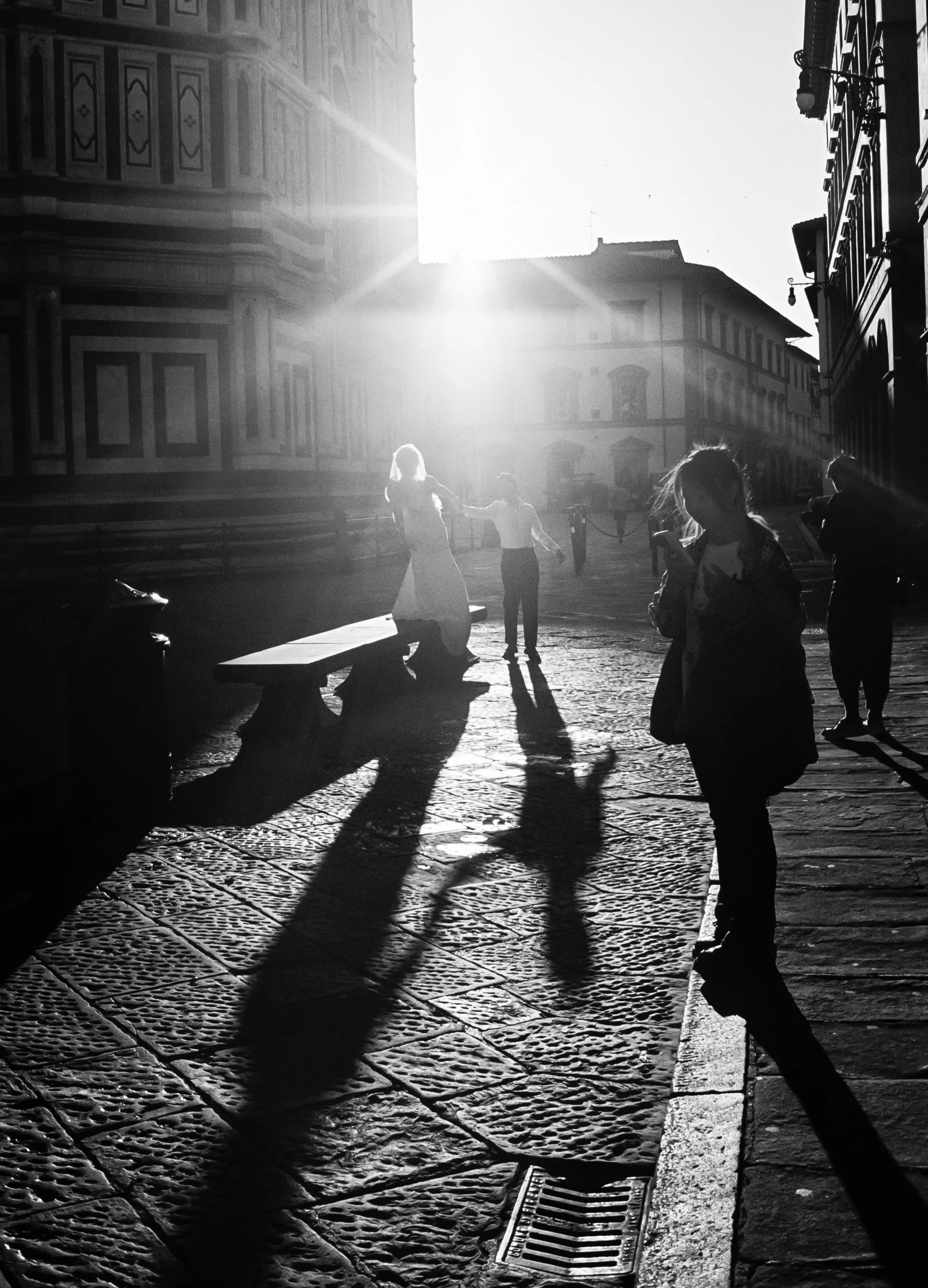 People walking and standing on a cobblestone street in Florence, Italy, with historic buildings in the background. The sun is setting, casting long shadows.