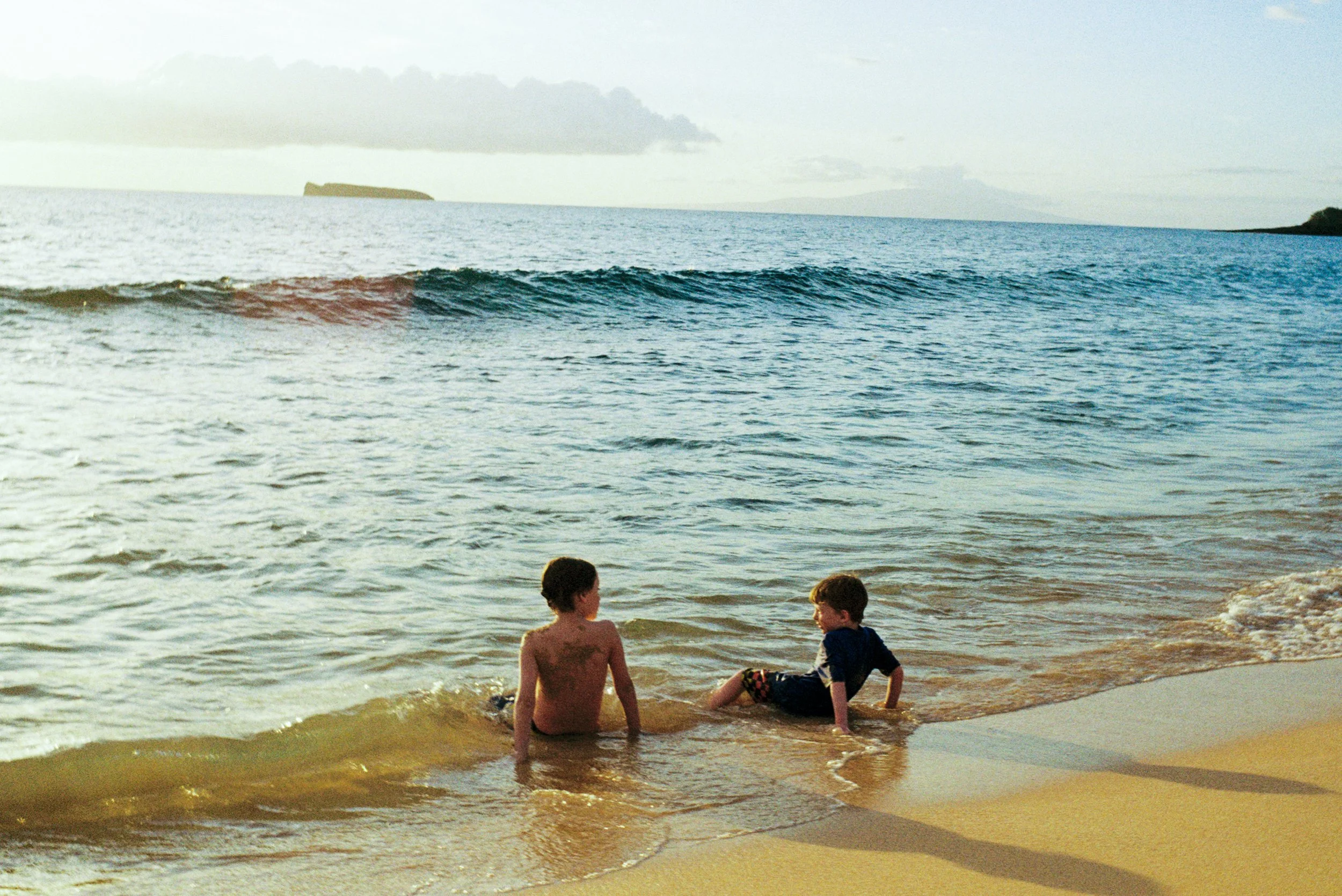 Two children sitting in shallow ocean water on a beach, with an island in the distance and a cloudy sky.