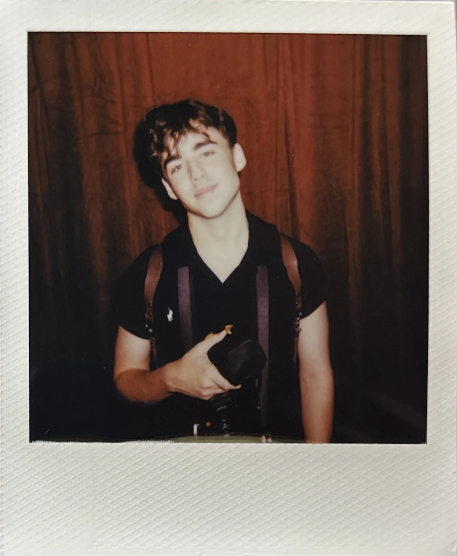 A young man with curly dark hair and light skin smiles slightly while holding a camera in front of a wooden wall.