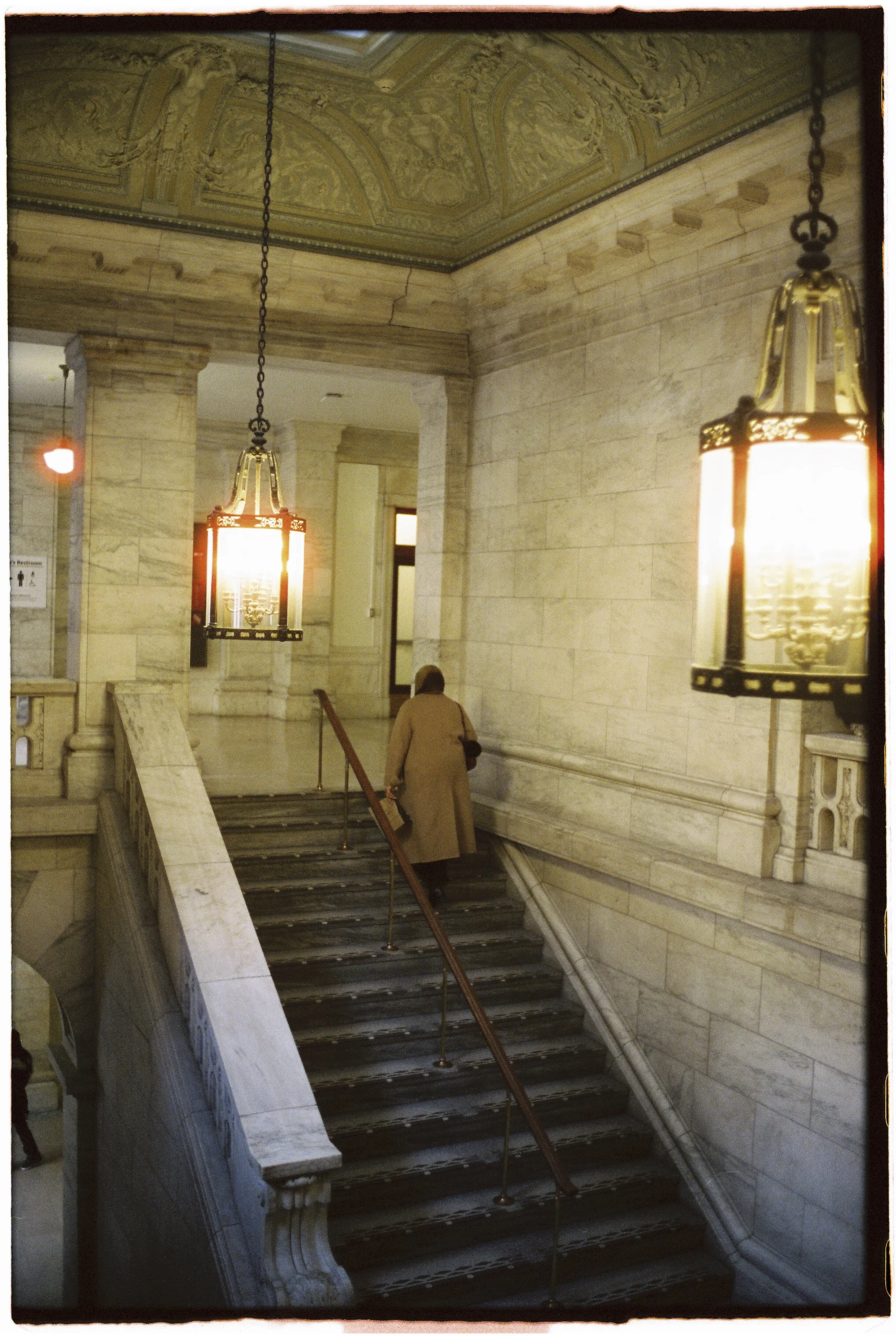 Interior view of a marble staircase in a historic building, with ornate ceiling details, hanging lantern-style lights, and a person wearing a tan coat ascending the stairs.