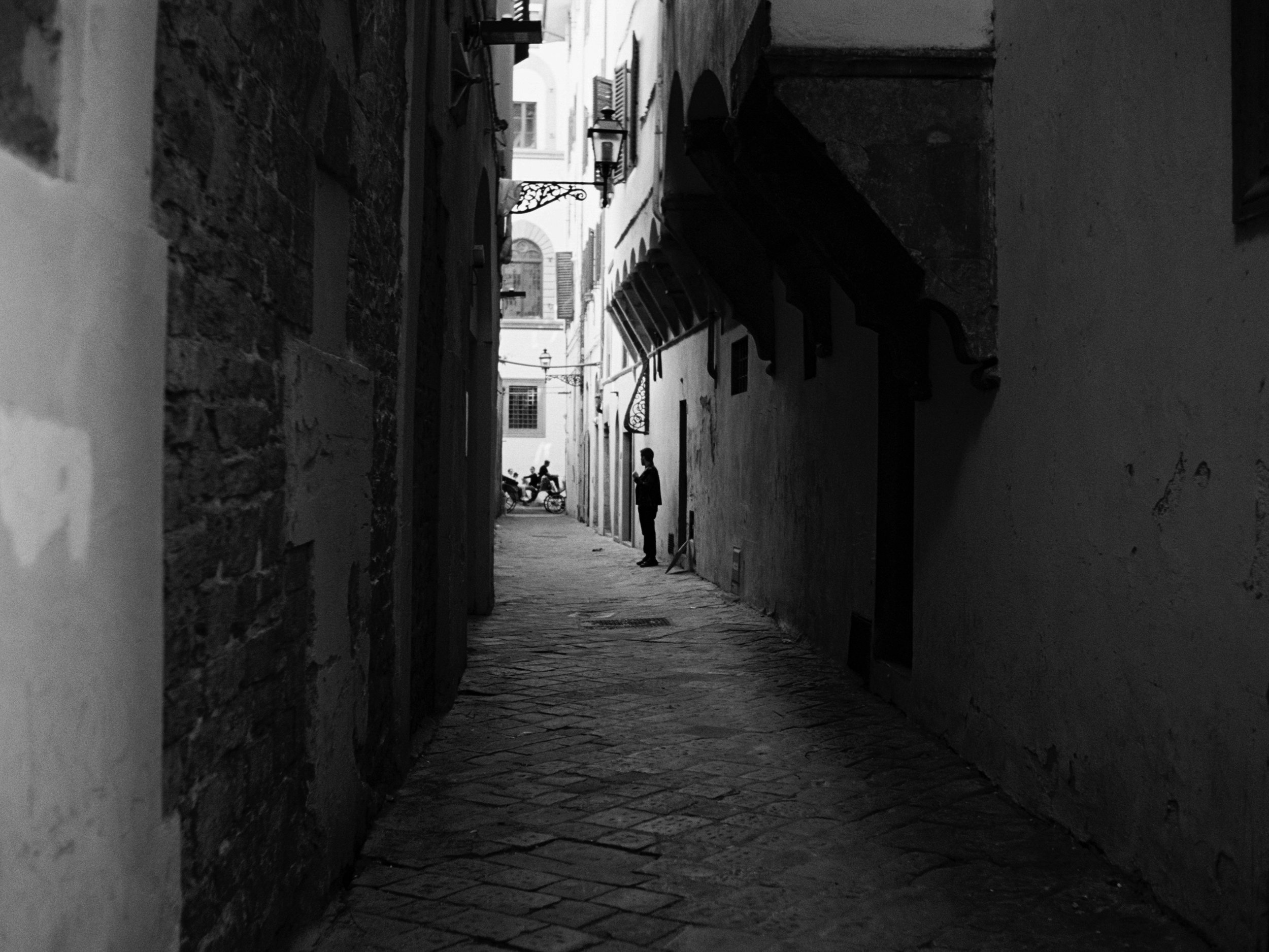 A narrow alleyway in a historic city with stone pavement, old stone and plaster buildings on each side, and a person standing and talking on the phone near the middle of the alley.