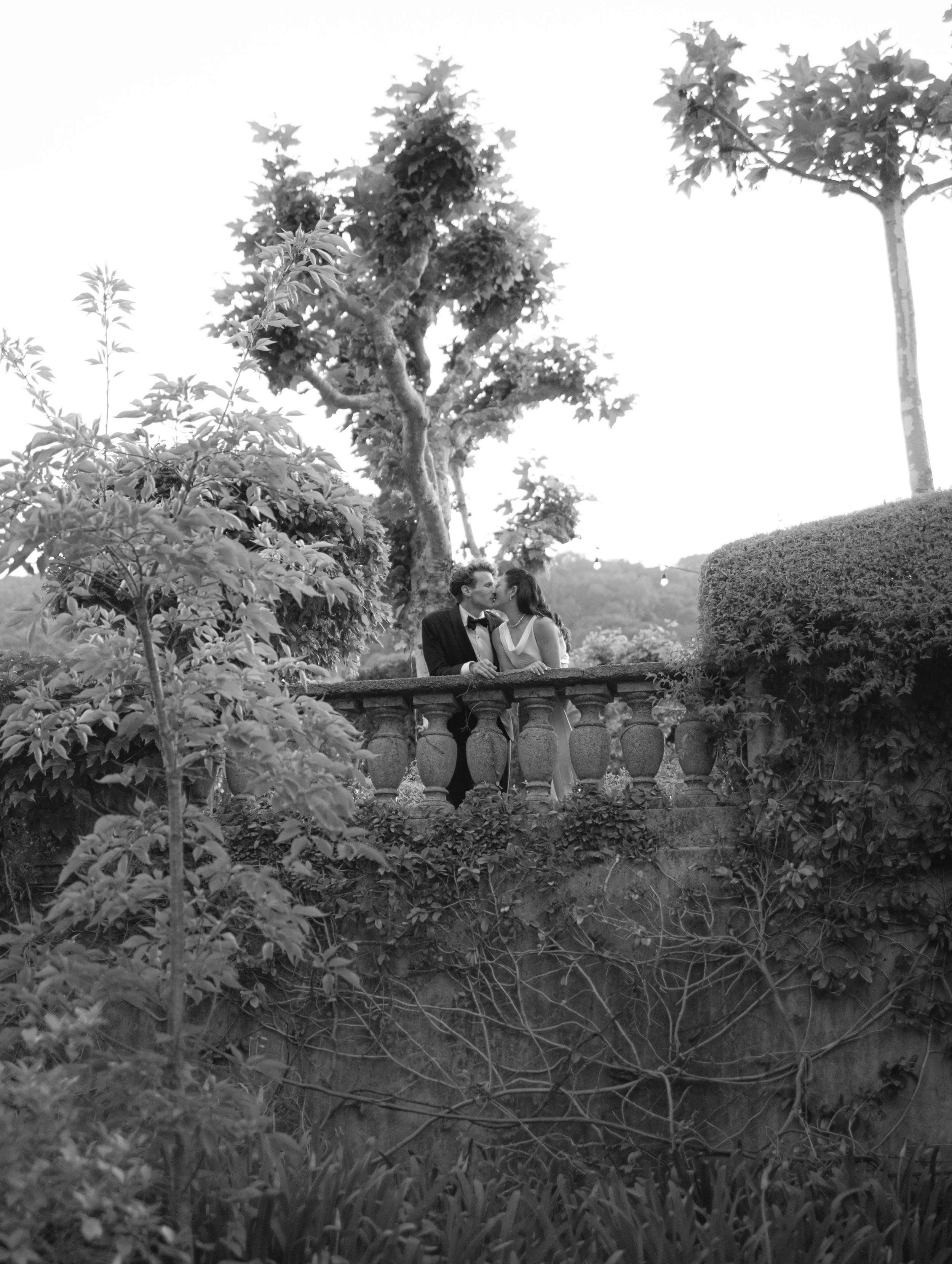 A black and white photo of a couple in wedding attire sharing a kiss outdoors, with trees and a stone railing in the background.