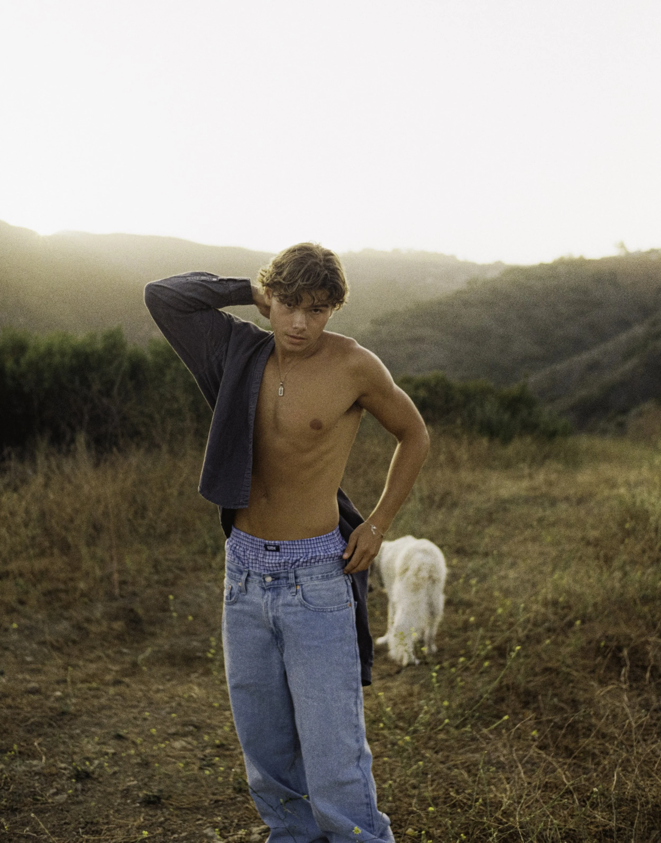 A young man with shaggy hair is standing outdoors on a hill, shirtless with a dark jacket hanging off his shoulders, wearing light blue jeans, with one hand on his hip and the other behind his head. There is a white dog in the background and rolling 