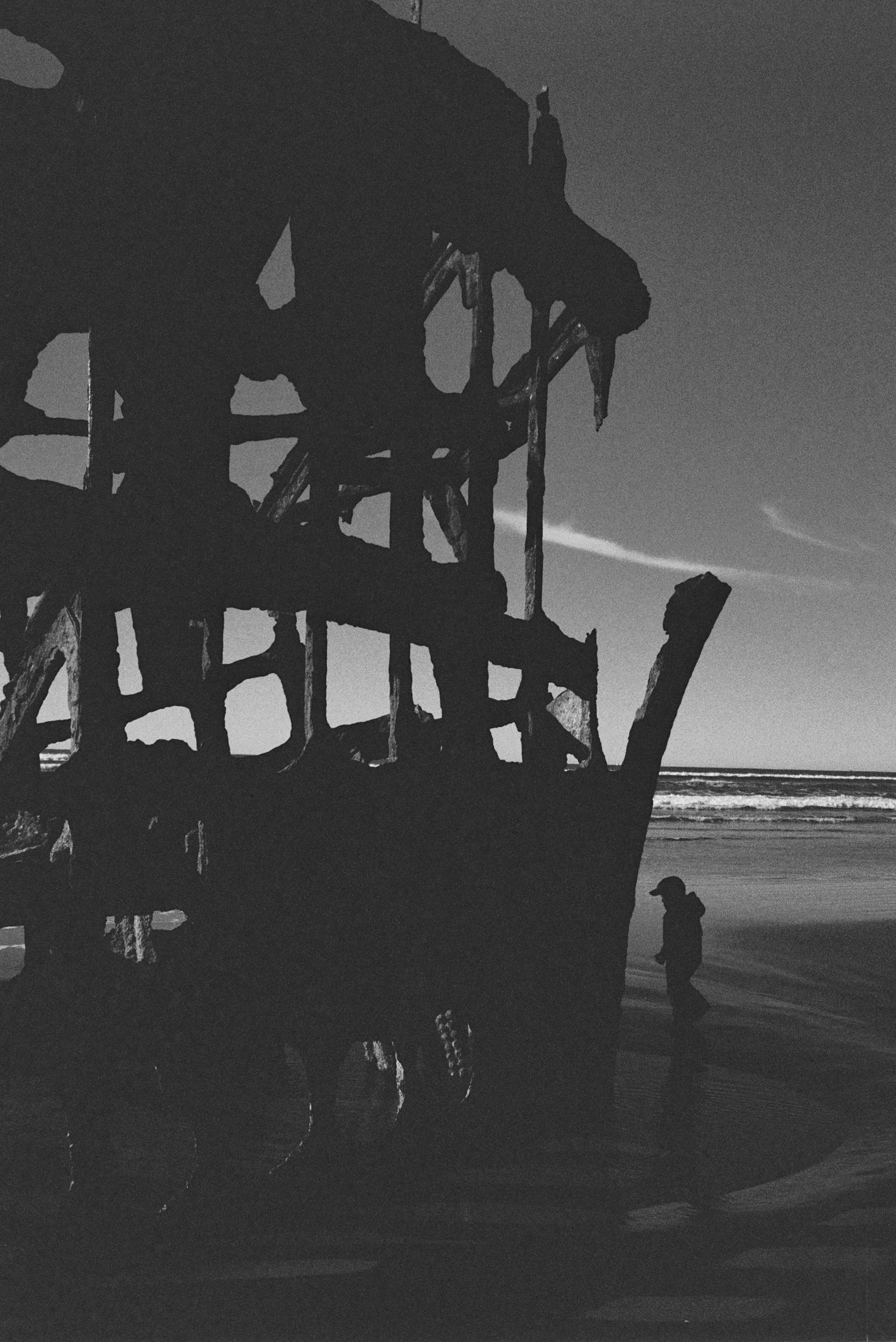 Silhouette of a person walking on a beach near a large, weathered wooden shipwreck with the ocean and sky in the background.