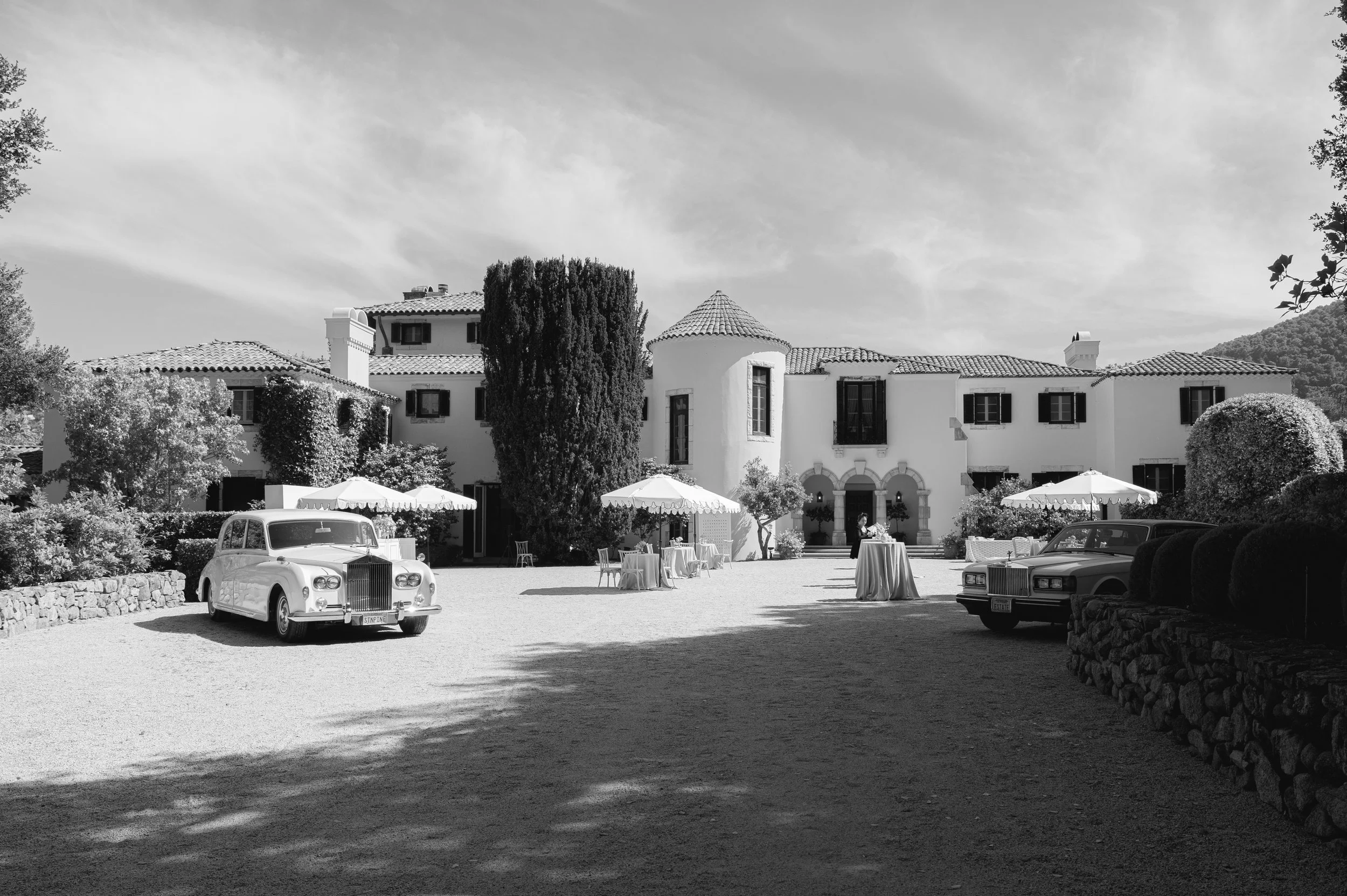 A black-and-white photo of a luxurious estate with a large house featuring a tiled roof and multiple stories, surrounded by trees, with vintage cars parked outside, and tables set for an outdoor event.