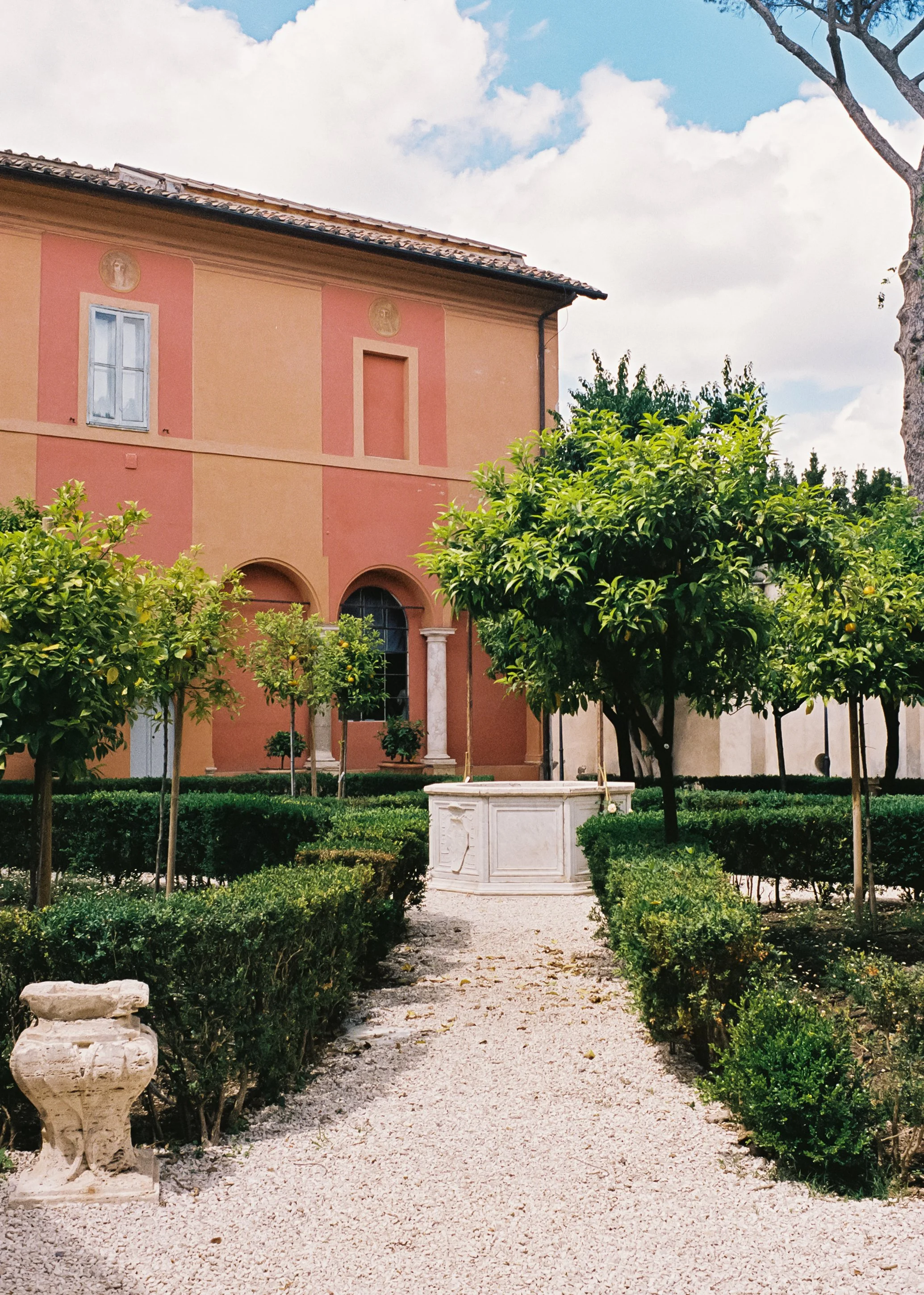 A garden with a gravel pathway, lush green trees, trimmed bushes, and a large building with pink and peach-colored walls in the background, under a partly cloudy sky.