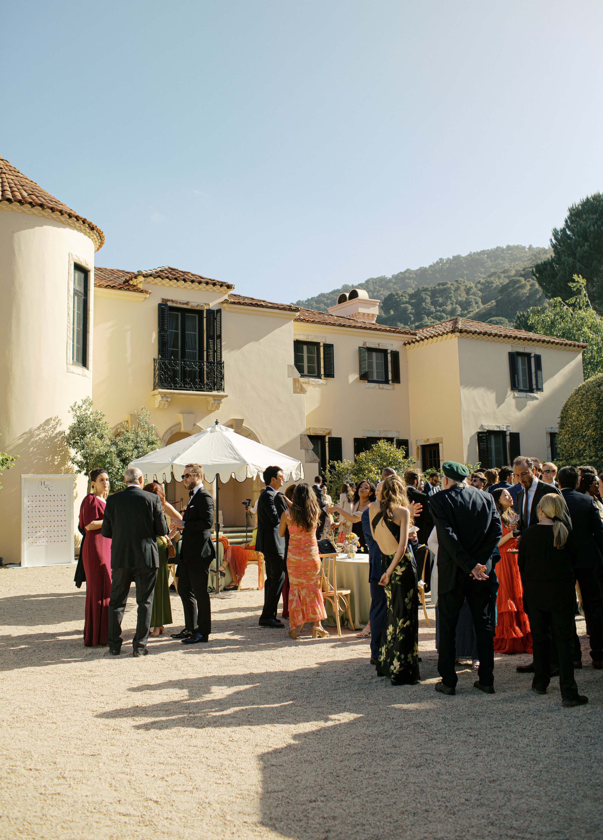 People gathered at an outdoor event at a vineyard estate, dressed in formal and semi-formal attire, with a large cream-colored house with black shutters and a terracotta tiled roof in the background, and mountains in the distance.