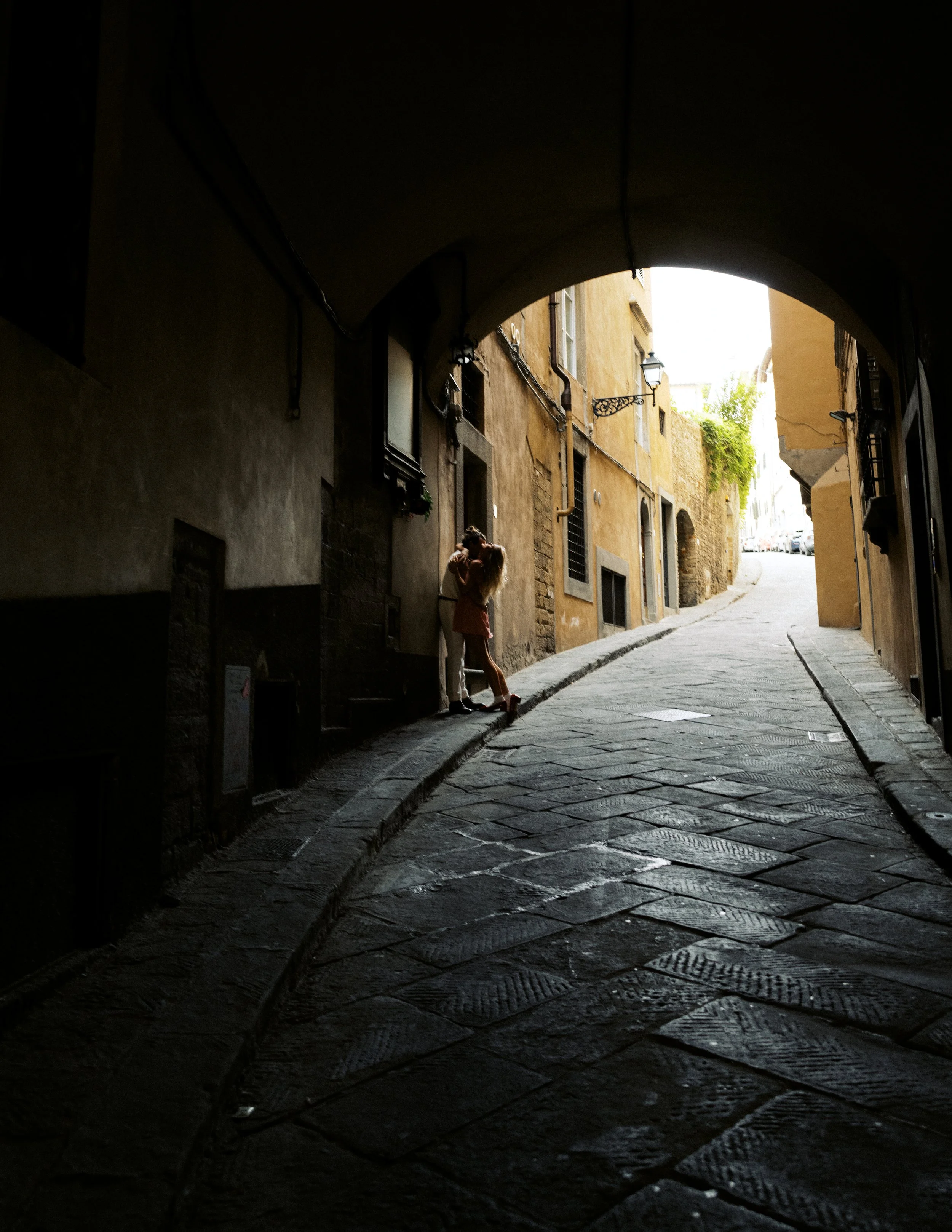 A narrow cobblestone street viewed from a dark archway, with a person standing near a building and holding a dog, sunlight illuminating the yellow and beige buildings at the end of the street.