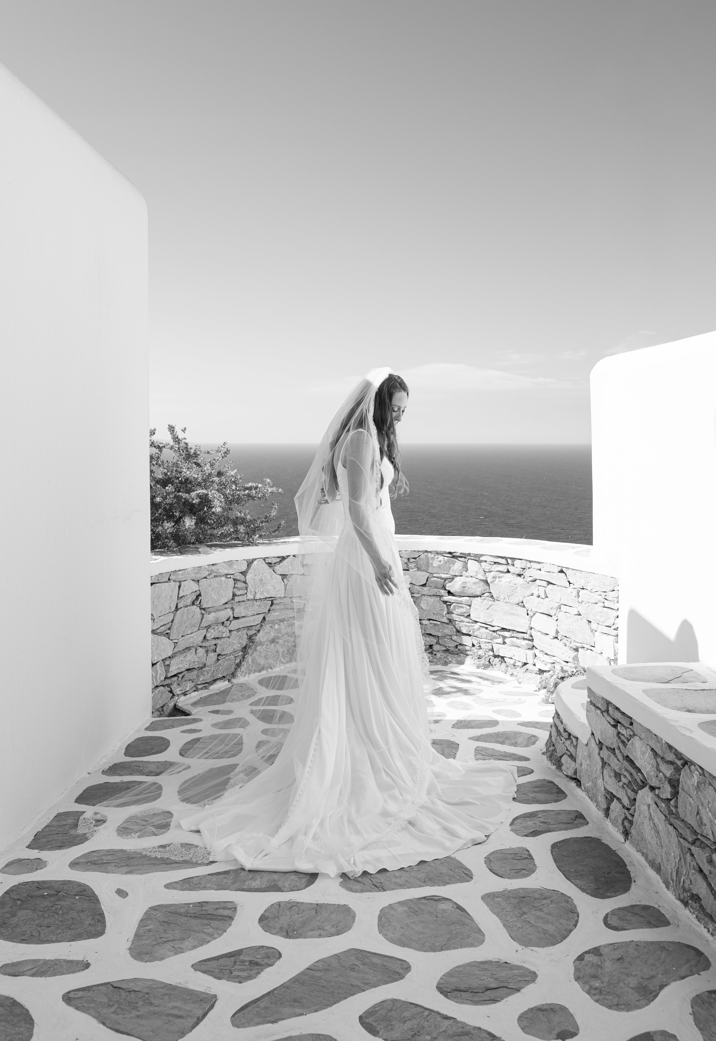 A woman in a wedding dress and veil standing outdoors on a stone patio overlooking the ocean, framed by white walls.