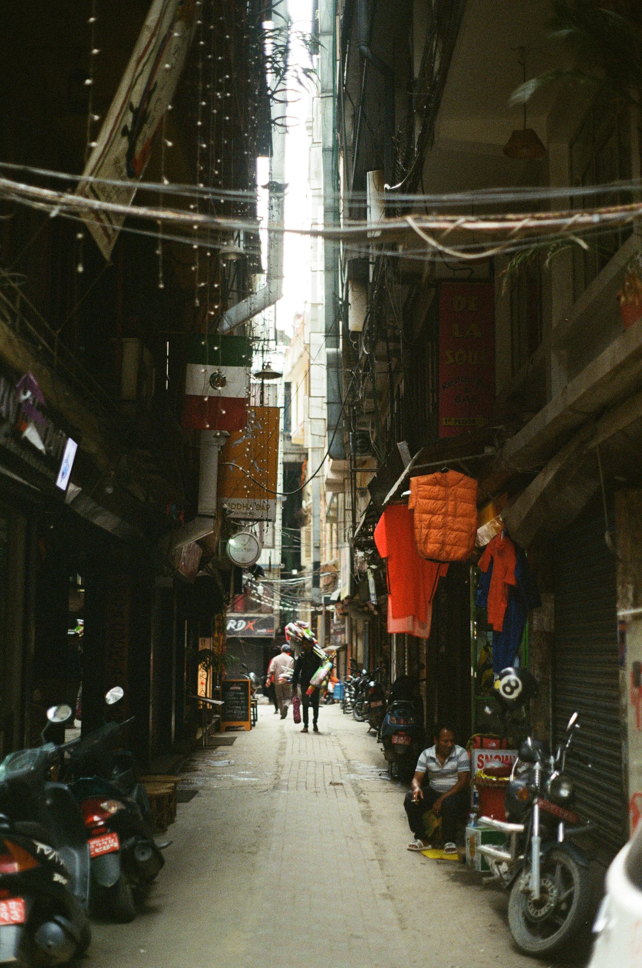 A narrow alleyway with parked scooters and motorcycles, with shops and hanging laundry on both sides, leading to a brighter street in the distance.