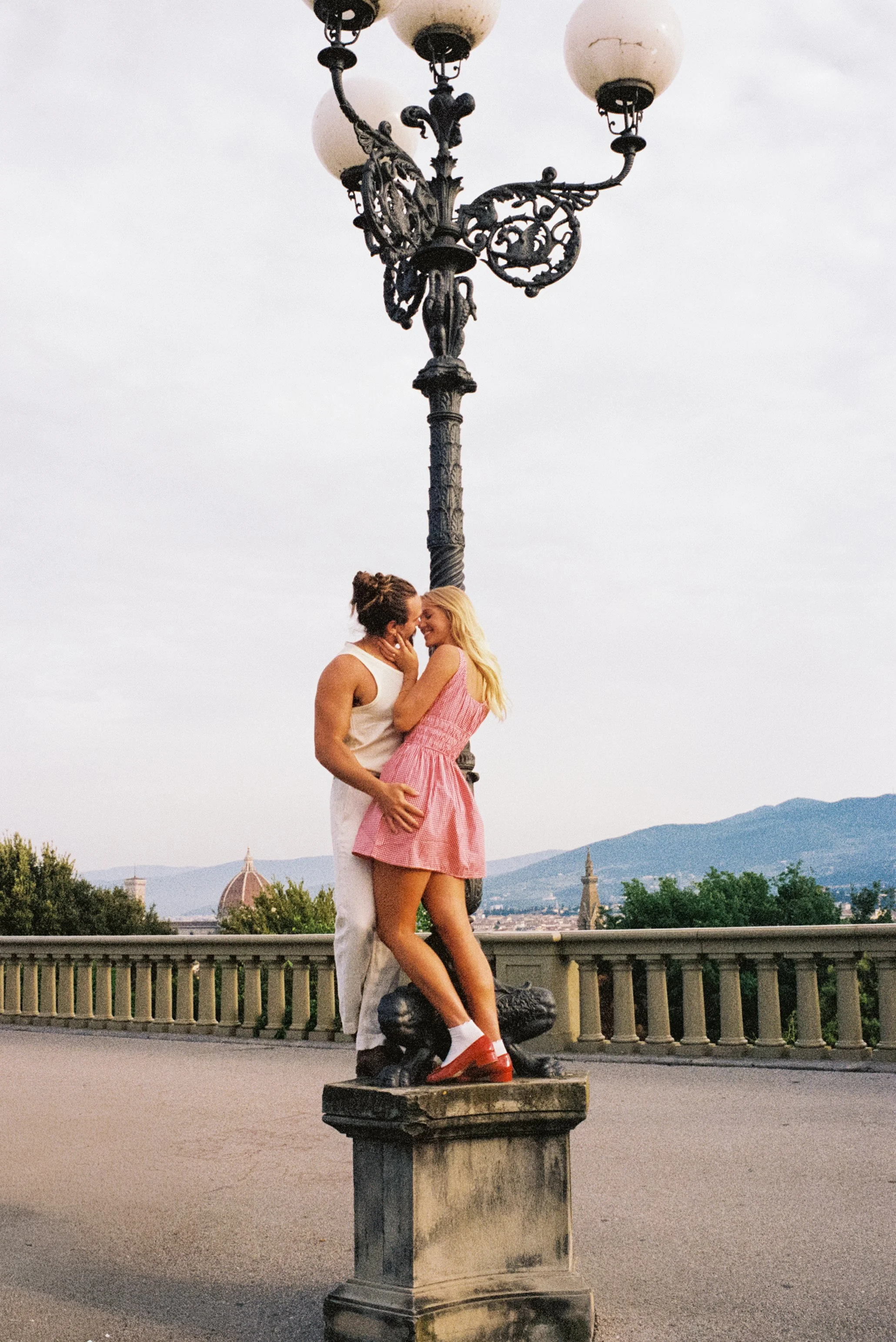 Two women embracing and smiling at each other next to a stone statue of a boy and a girl on a pedestal during daytime. A decorative street lamp is above them, and a cityscape with mountains and historical buildings is in the background.