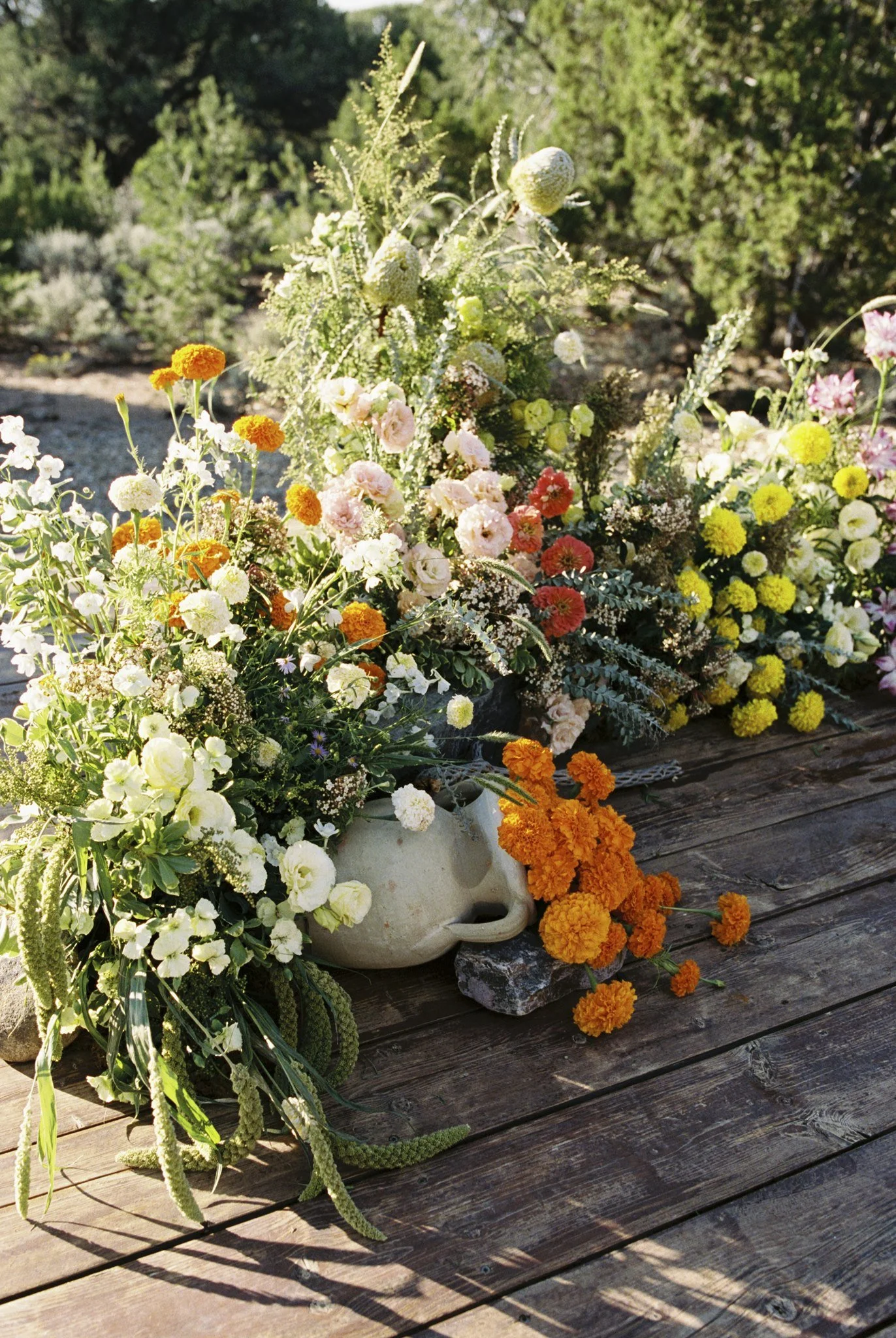 Various colorful flowers, including marigolds, petunias, and daisies, arranged in white ceramic and black stone containers on a weathered wooden surface outdoors, with a backdrop of green bushes and trees.