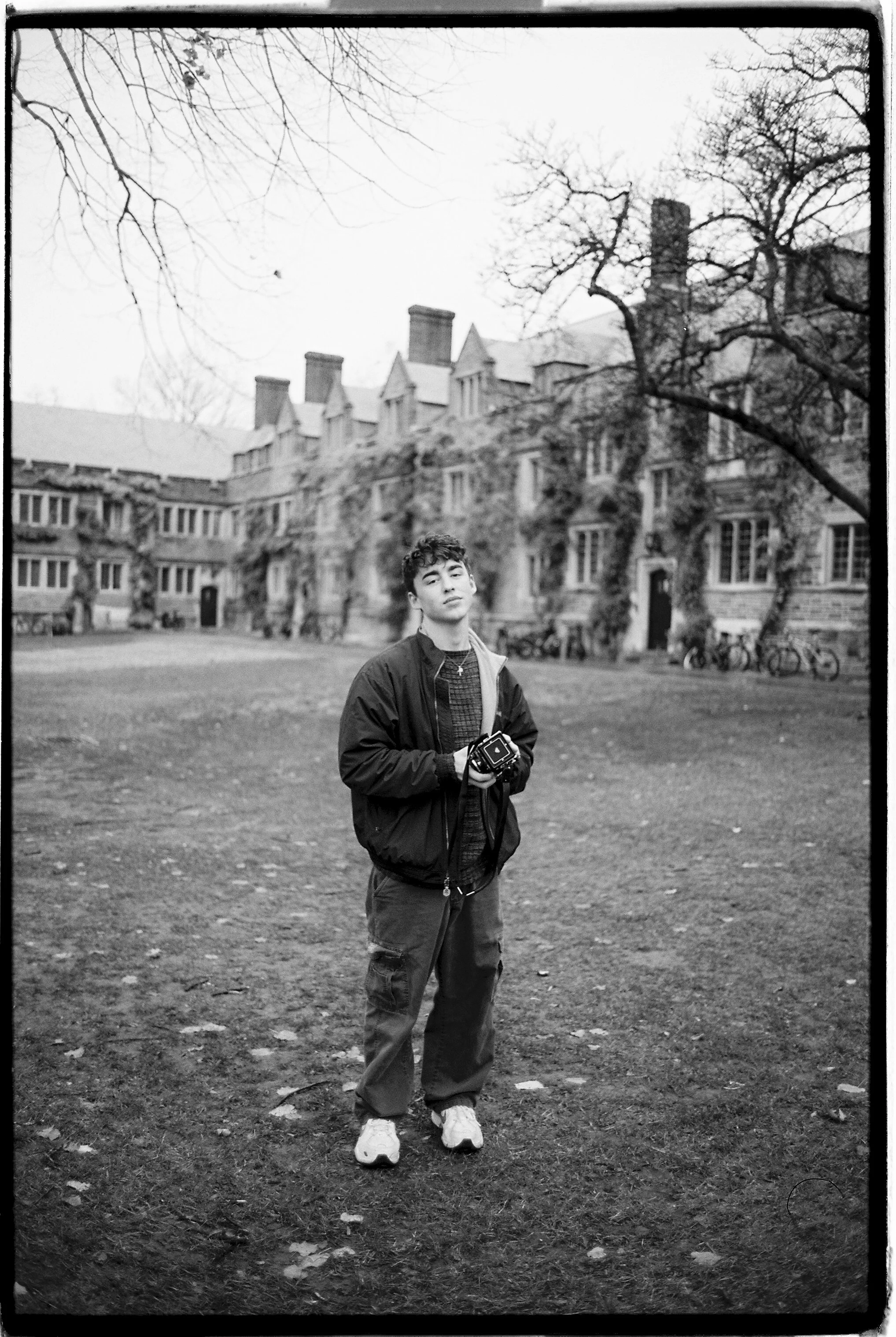 A young man standing on a grassy area holding a camera, with leafless trees and historic buildings in the background, black and white photo.