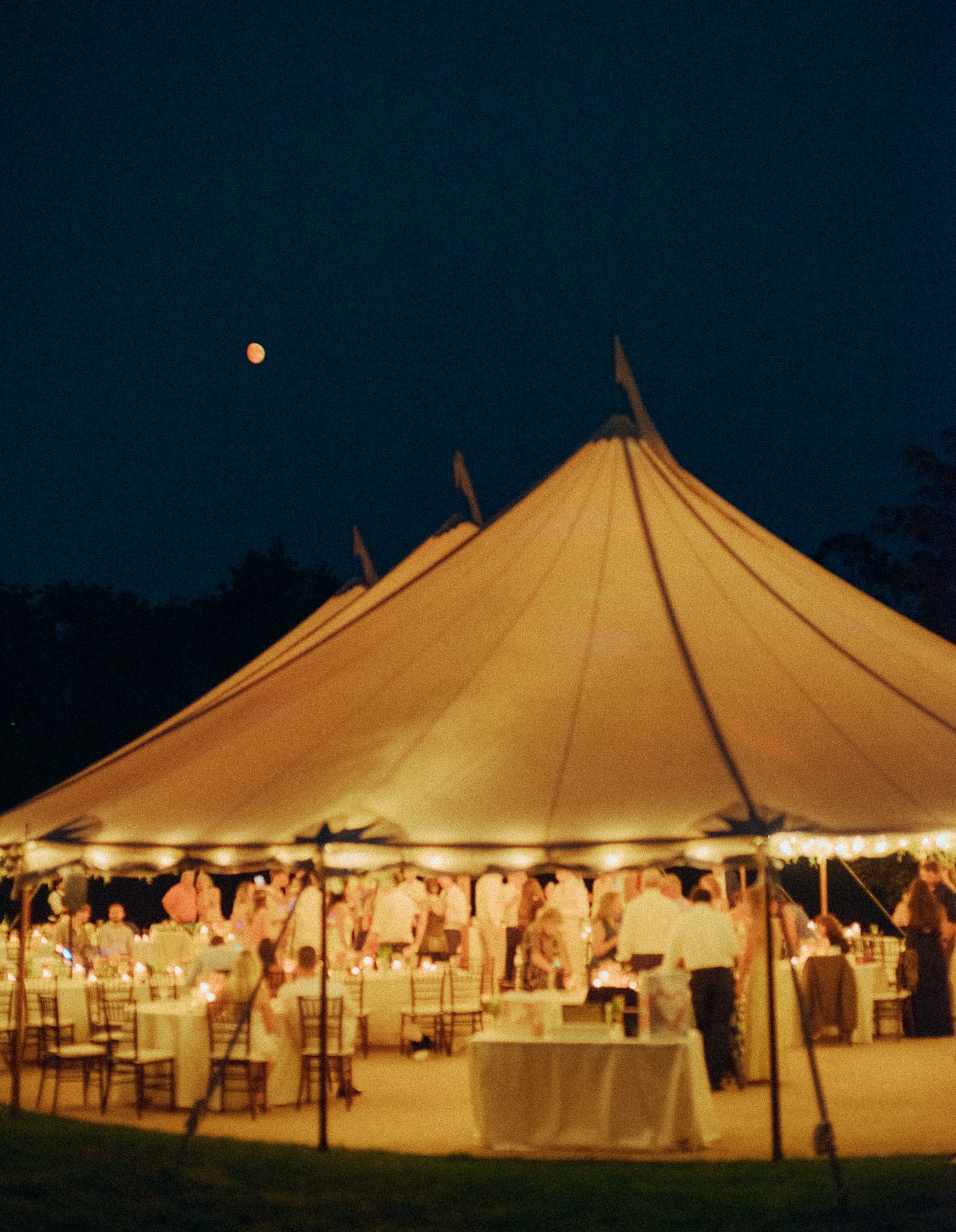 Night scene with a large white event tent, string lights inside, and people gathered underneath, with a view of the moon in the dark sky.