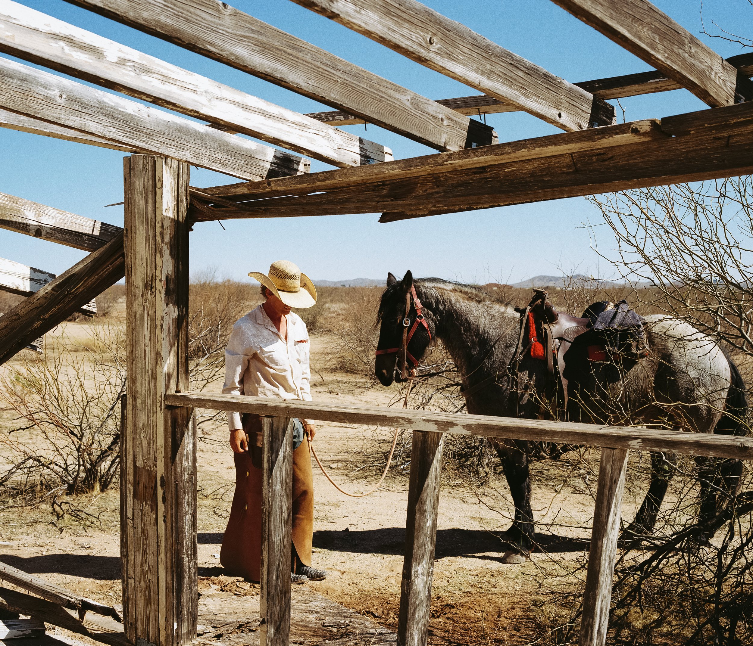 A person wearing a cowboy hat, white shirt, and brown skirt standing next to a gray horse with a red saddle and bridle, behind weathered wooden fence in desert landscape with sparse bushes and distant mountains.