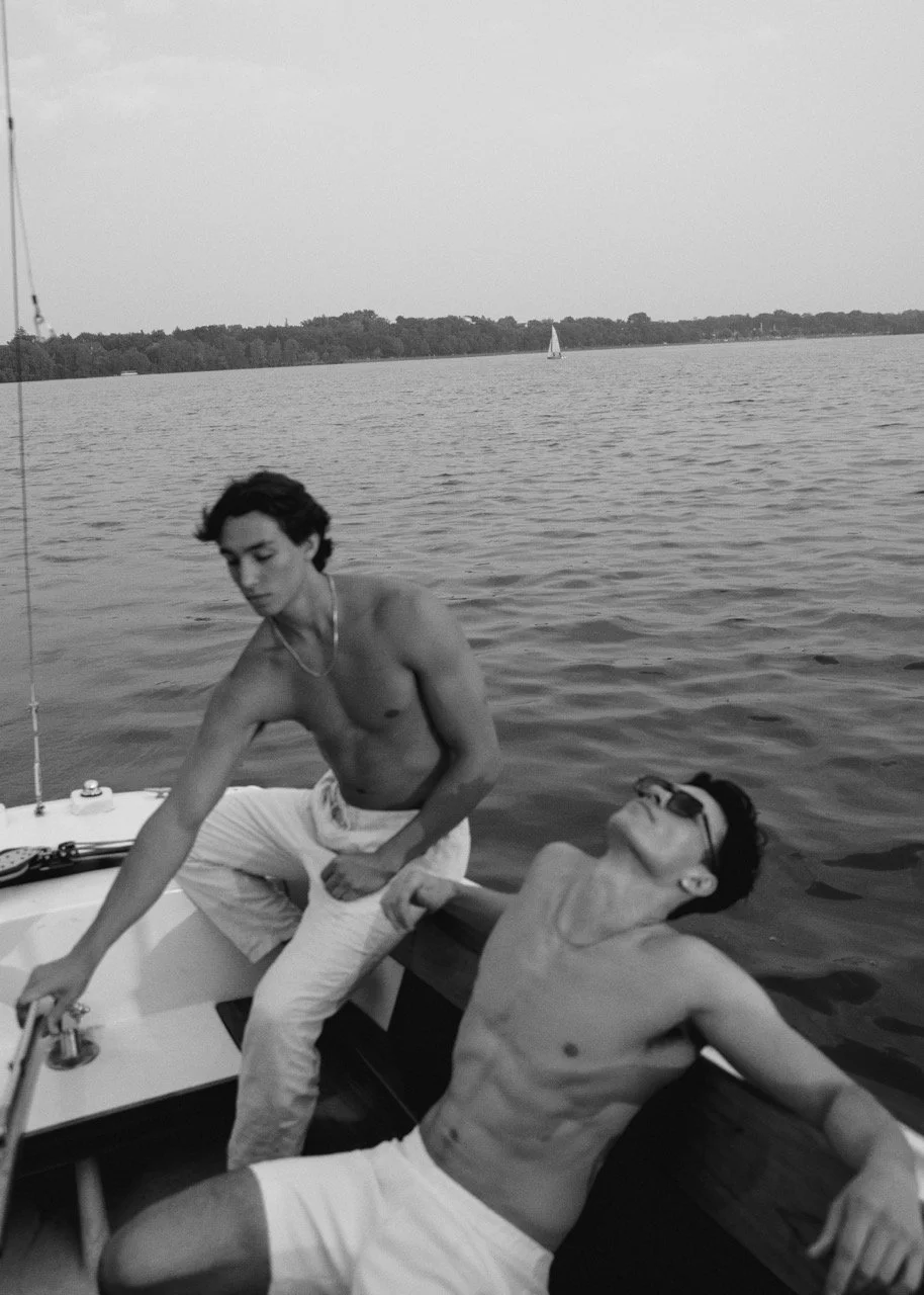 Two shirtless young men on a boat, one sitting and the other leaning back, with a lake and a sailboat in the background.