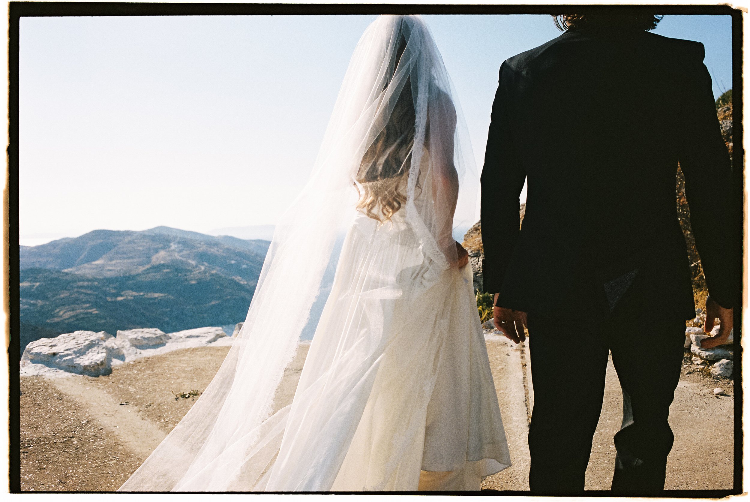 A bride in a wedding dress with a veil and a groom in a black suit holding hands, standing outdoors on a rocky area with mountains in the background