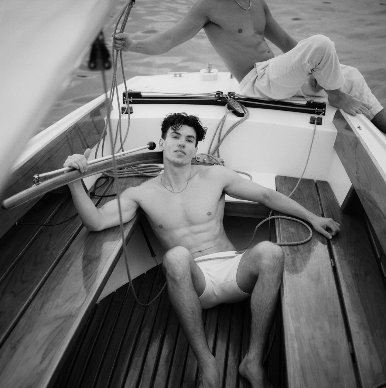 Two young men relaxing on a sailboat, shirtless, wearing shorts, with water in the background, in black and white.