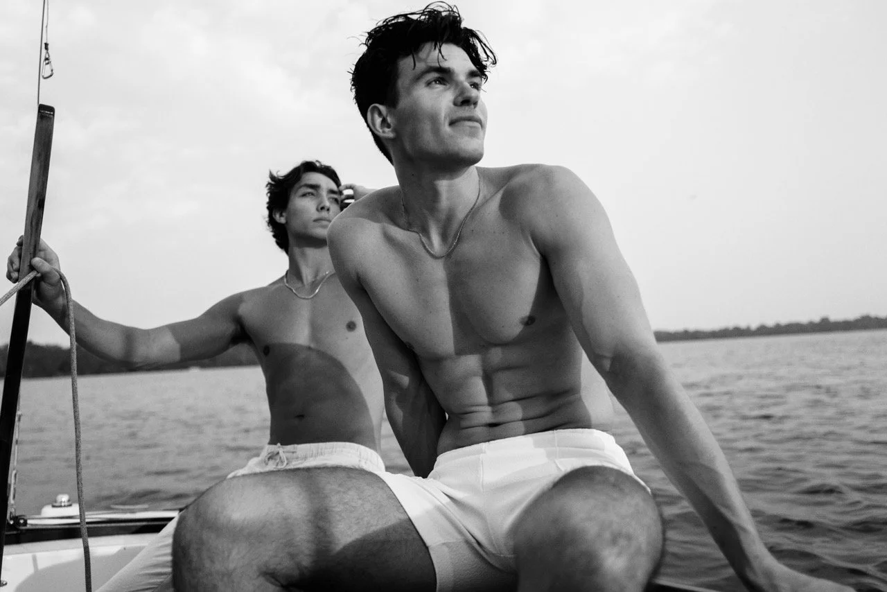 Two shirtless men on a boat looking off into the distance on a body of water