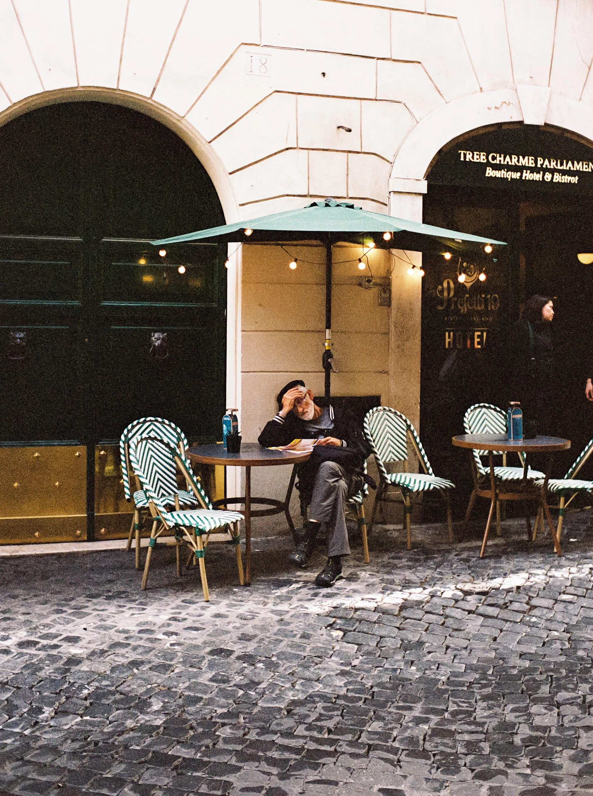 A person sitting alone at a small outdoor table outside a cafe under a string of lights, with a white and green striped chair, in front of a building with a sign for Tree Charme Parliament, a boutique hotel and bistro.