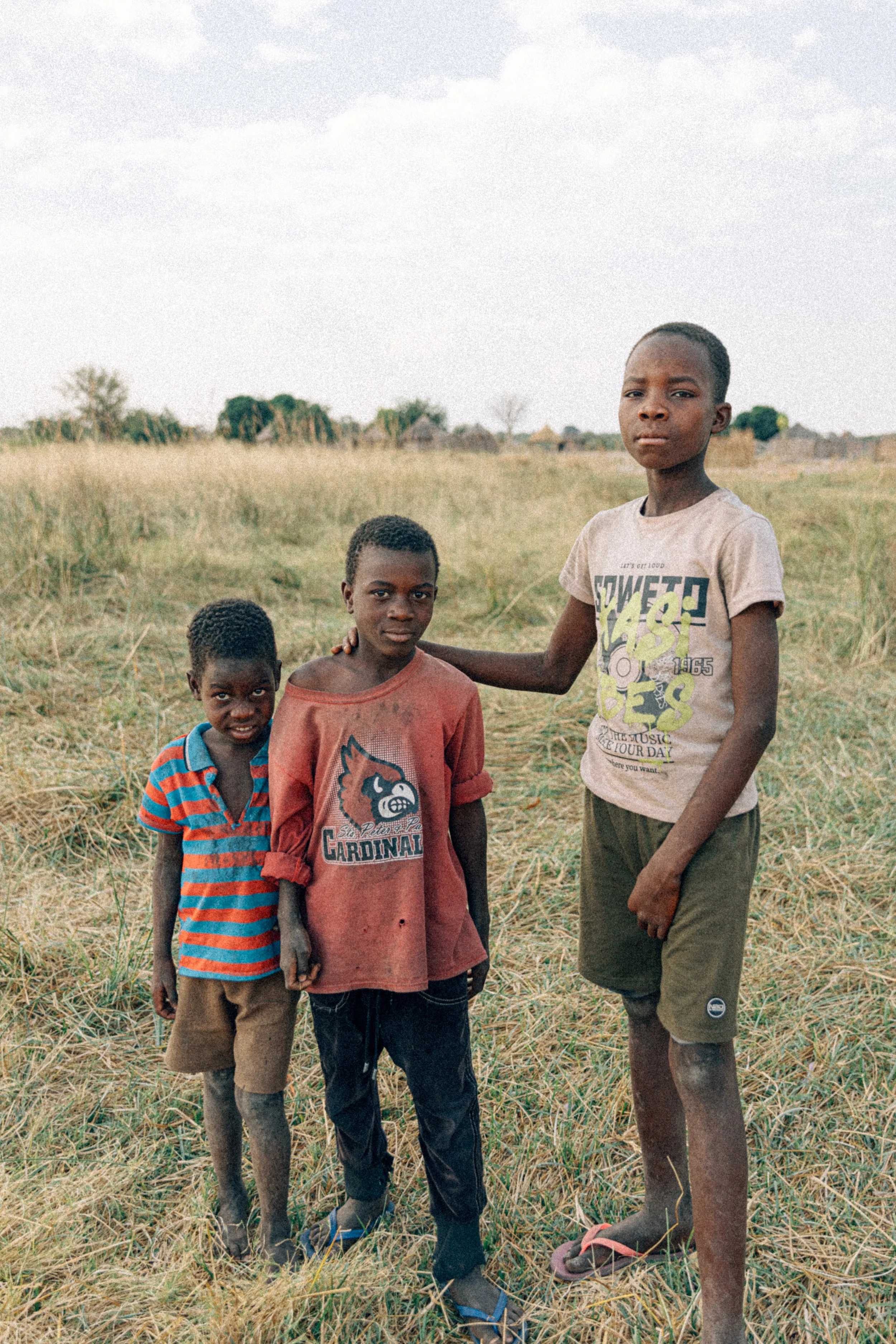 Three young boys standing outdoors in a grassy field, with small huts and trees in the background. One boy has his arm around another's shoulder.